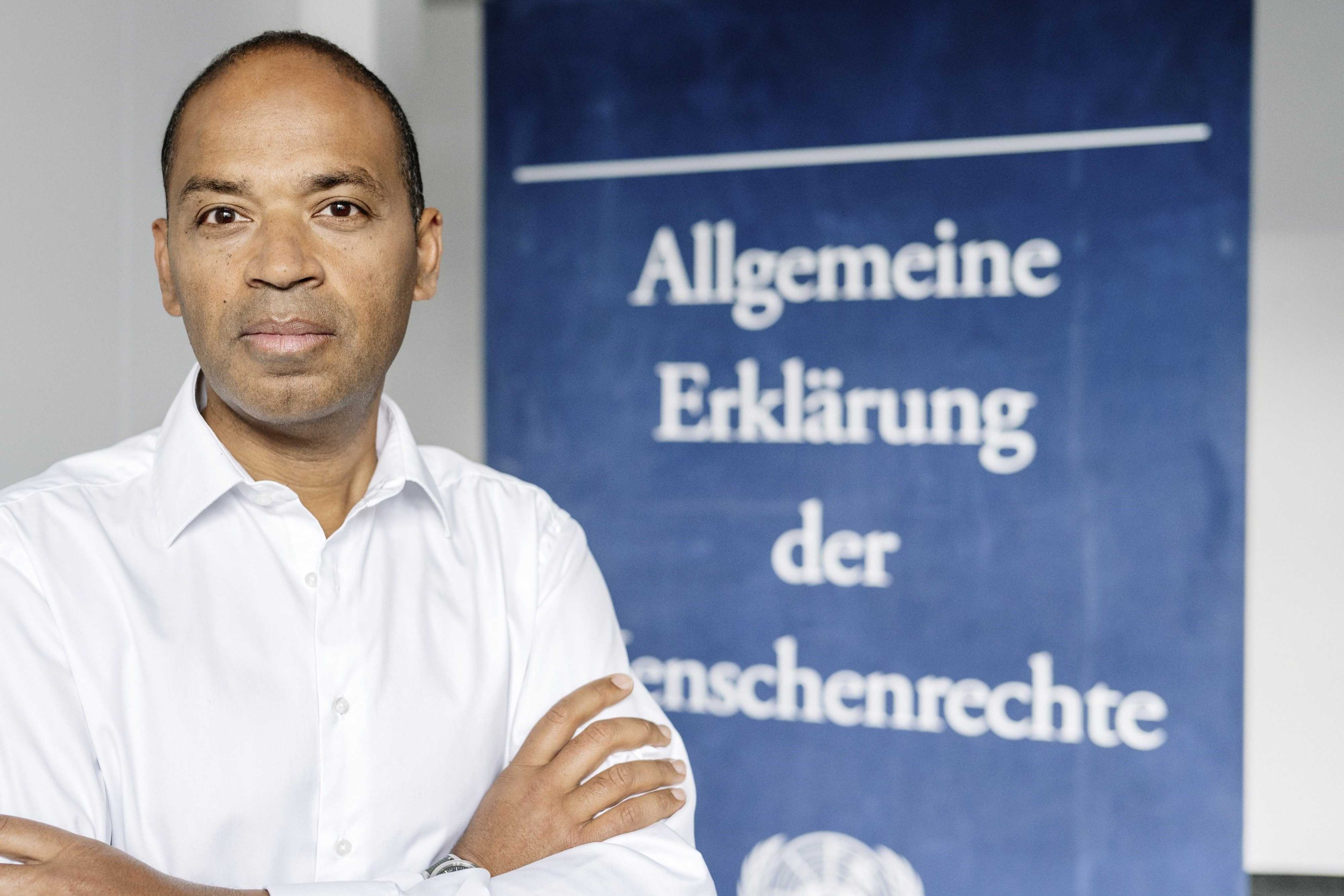 Markus Beeko is standing in front of the Declaration of Human Rights as a poster and looks into the camera