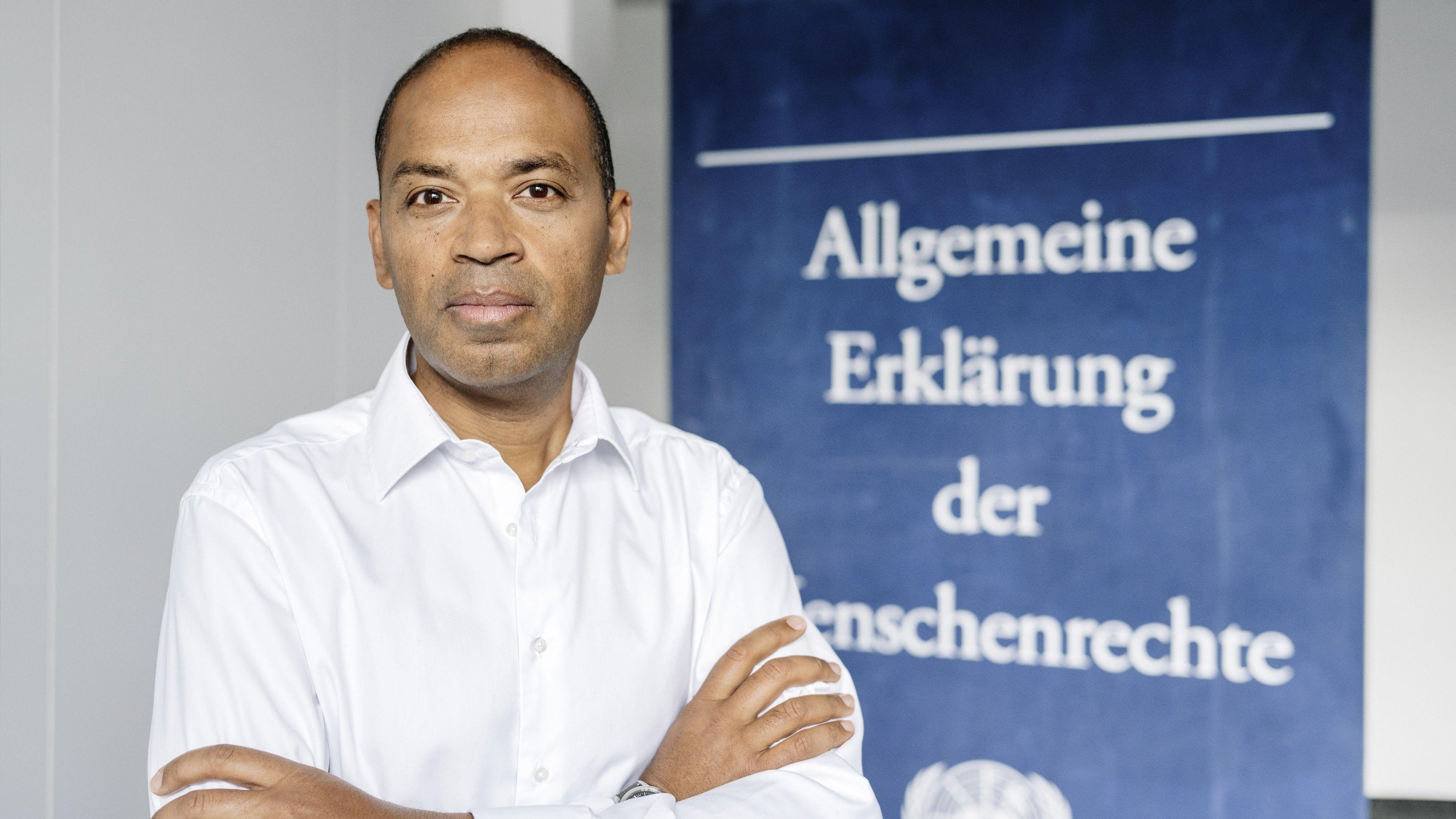 Markus Beeko is standing in front of the Declaration of Human Rights as a poster and looks into the camera