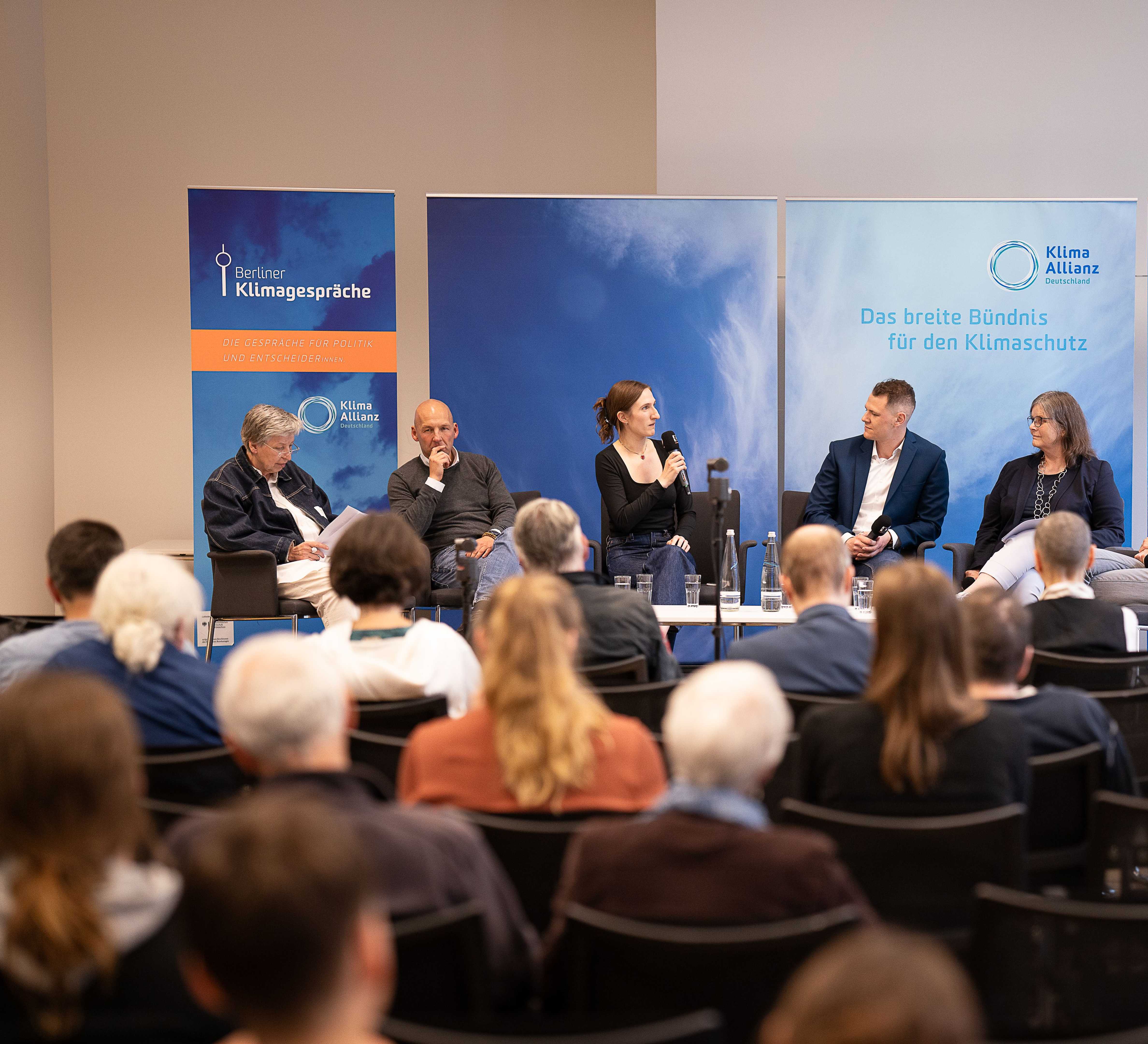 Six people are sitting on a panel and are talking to each other. People are sitting in the room and are listening.