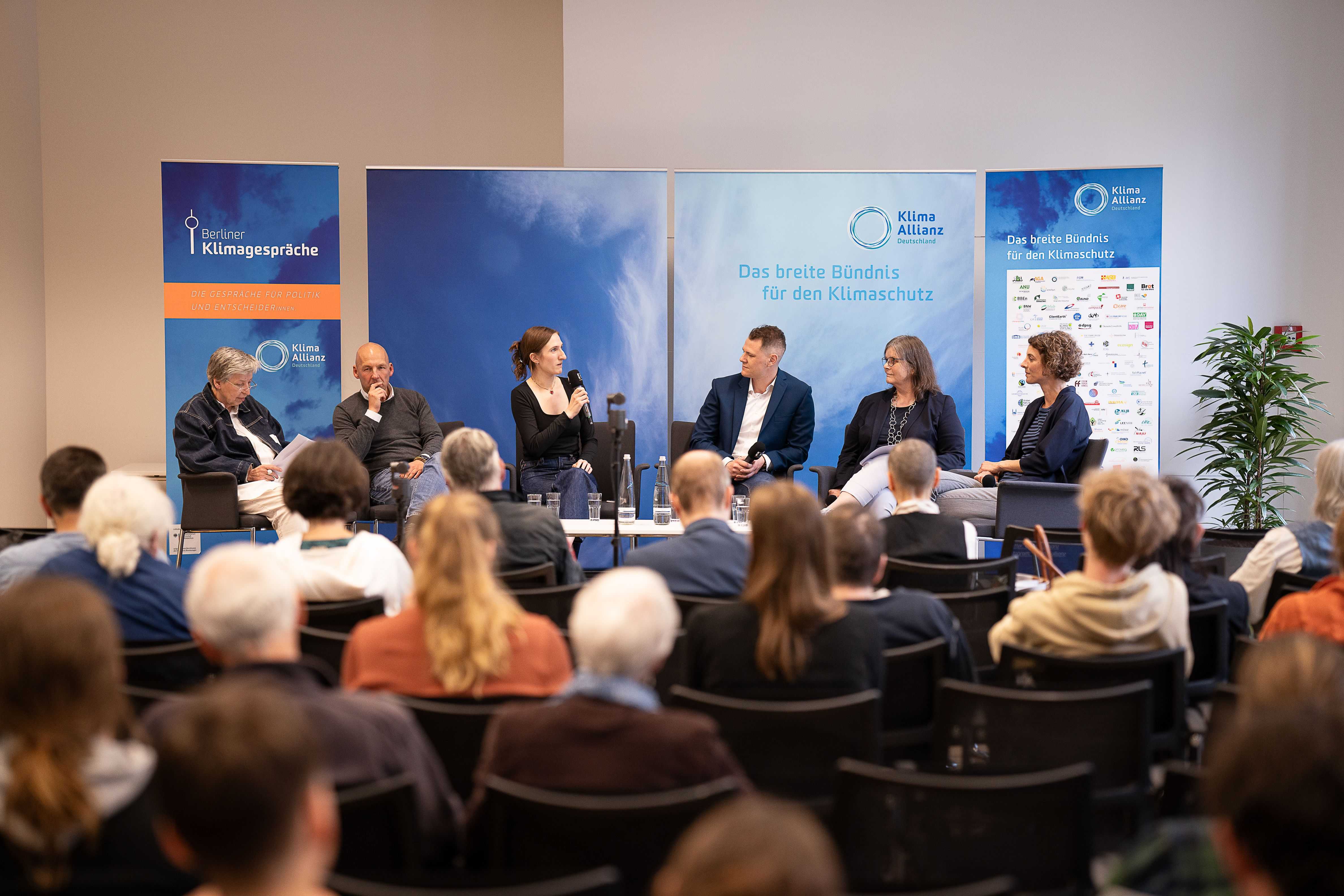 Six people are sitting on a panel and are talking to each other. People are sitting in the room and are listening.