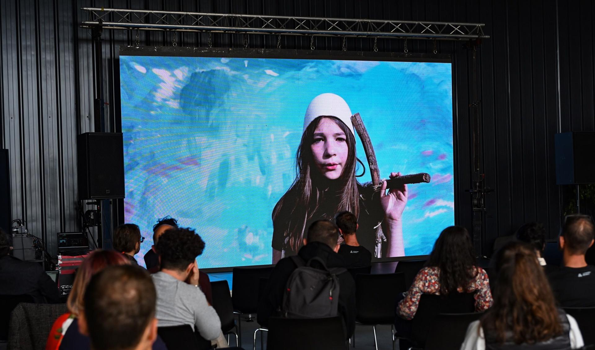 A screen is showing a woman and in front of the screen are many people sitting and watching.