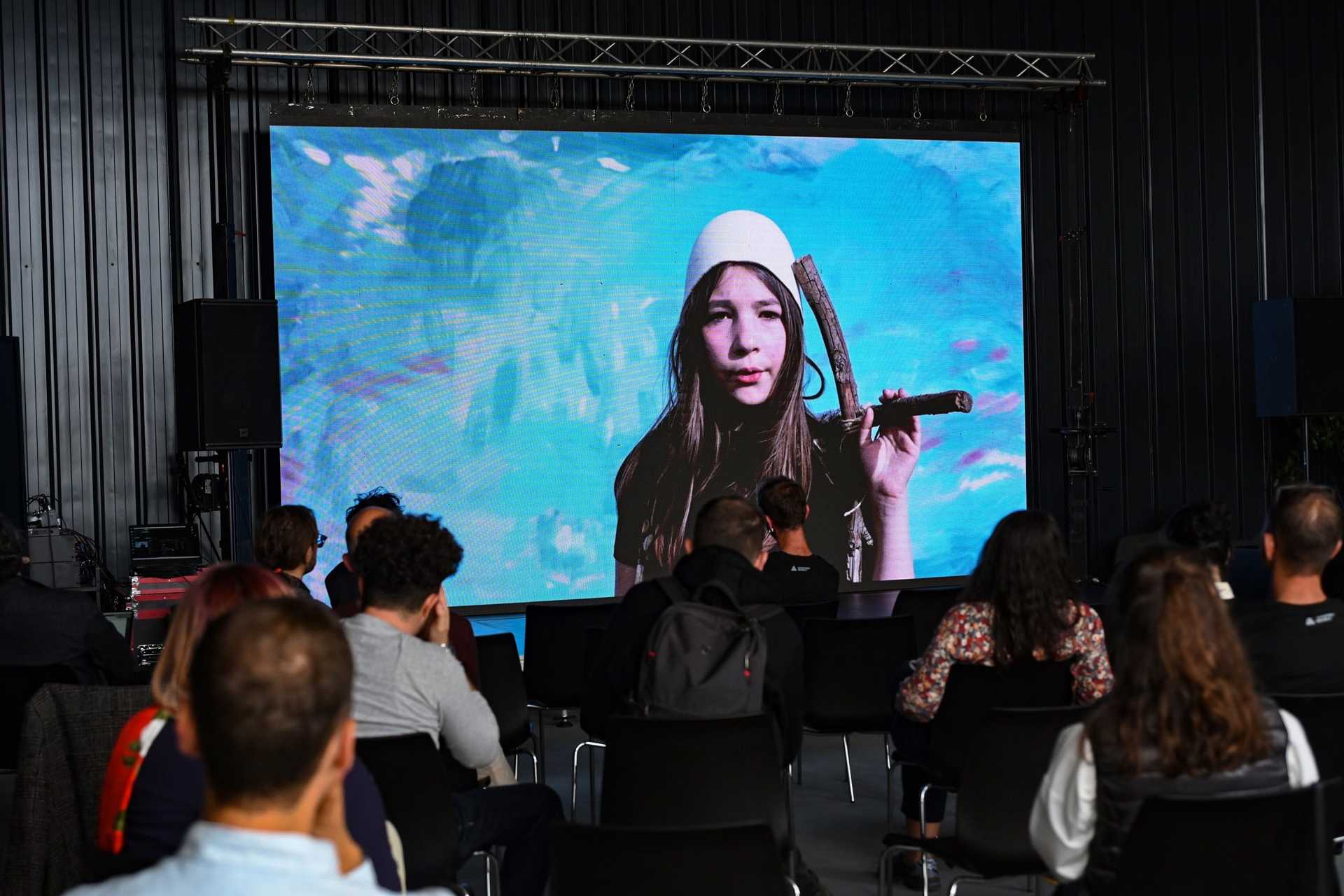A screen is showing a woman and in front of the screen are many people sitting and watching.