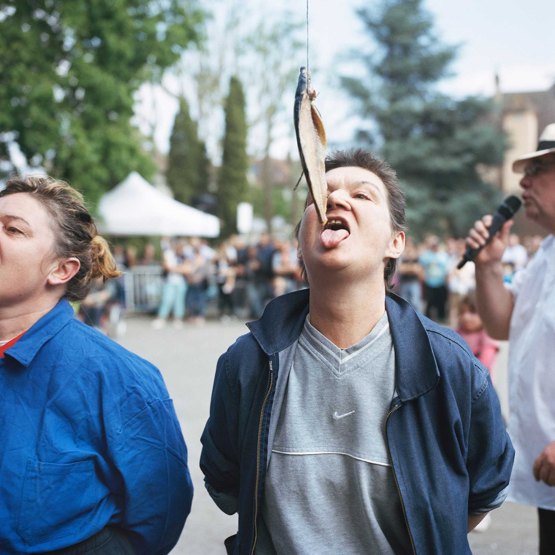 A person has her hands on the back and is trying to eat a fish on a stick and a rope only with the mouth. Next to the person is another person, doing the same. 