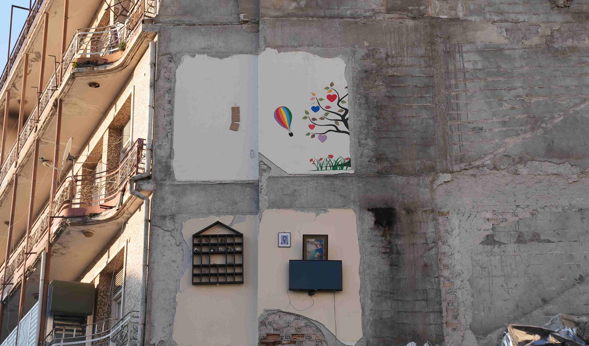 A wall of a demolished house. There are leftovers of the walls of the rooms, one where still a TV-screen is attached to the wall, another one with a shelf.