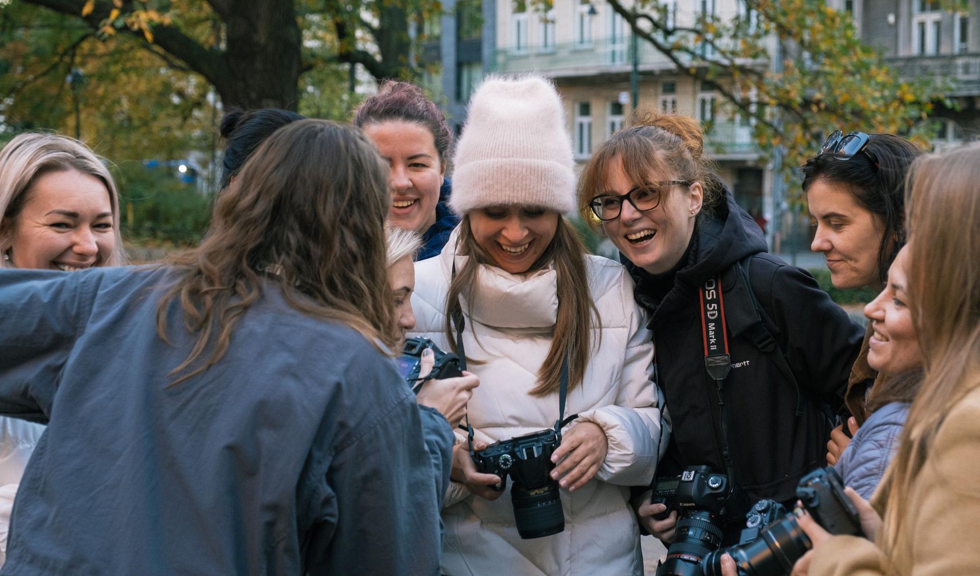 A group of women and girls gathered and are looking at a camera to see pictures one of them made. They are laughing.