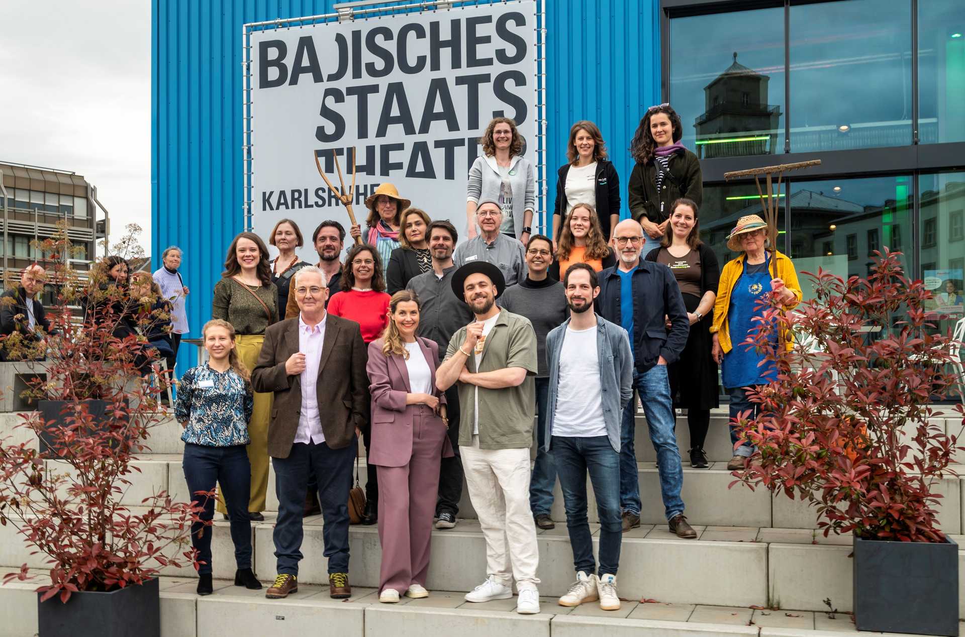 Eine Gruppe von Menschen steht auf einer Treppe aus Beton vor einem blauen Gebäude und schaut in die Kaera