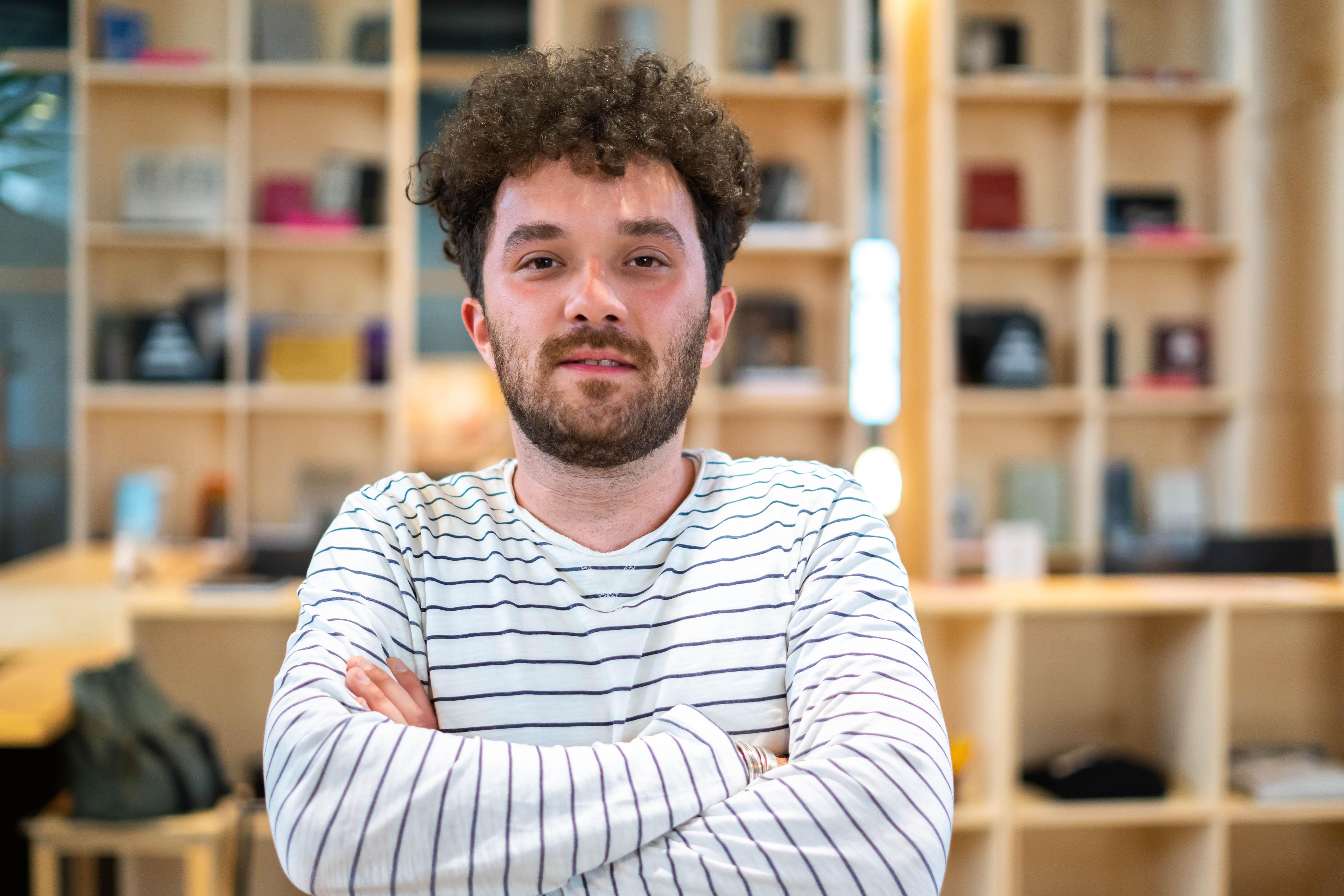 Dion Zeqiri is standing in a room in the Autostrada Hangar in front of a shelve with many items in it. Dion smiles into the camera, folds his arms, has short curly hair and wears a black and white striped shirt.