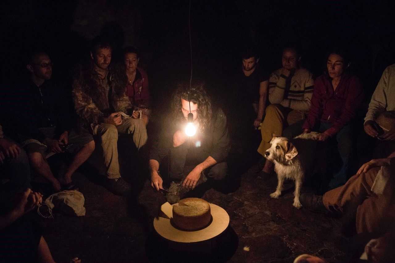 People are sitting around a cheese. One person sits in the middle and cuts the cheese. There is one dimmed light shining.