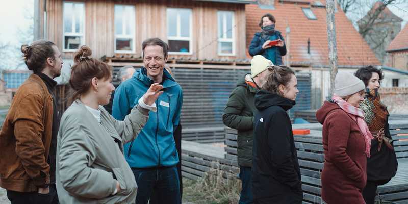 People are standing outside on a construction side in a smaller town oder village. They are talking to each other and are laughing.