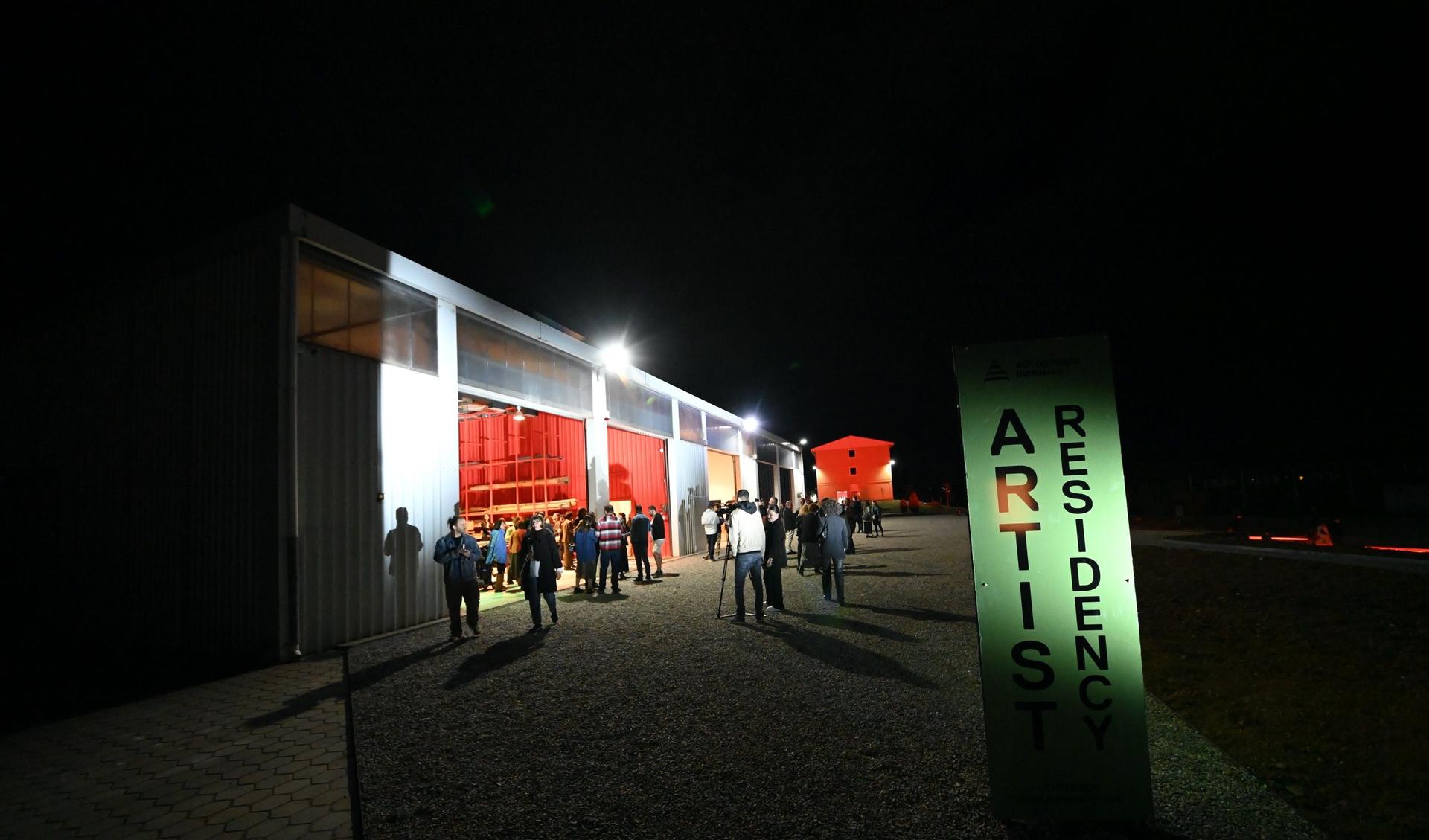 A hangar is in the night with open doors and many people walking around. 