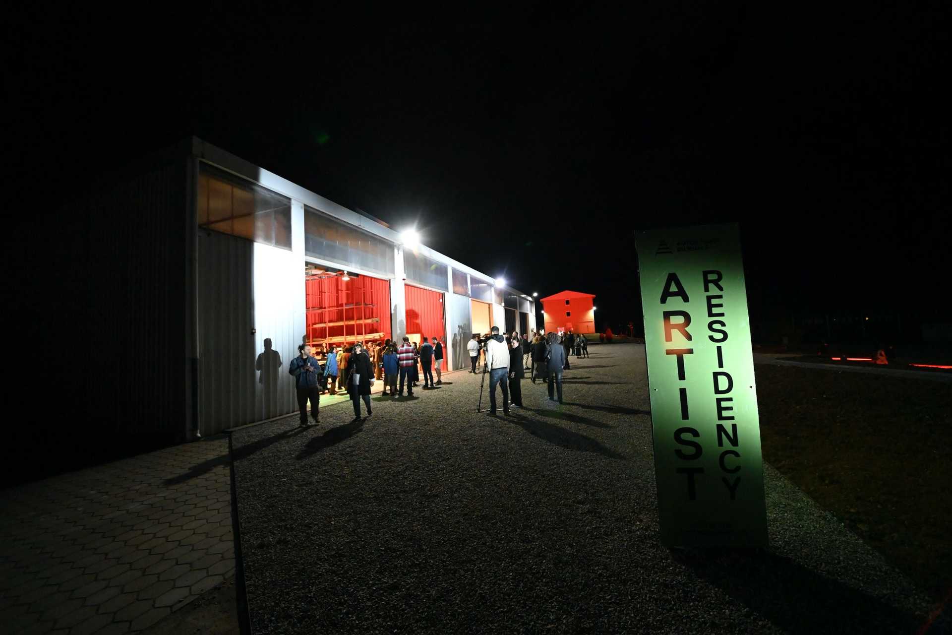 A hangar is in the night with open doors and many people walking around. 