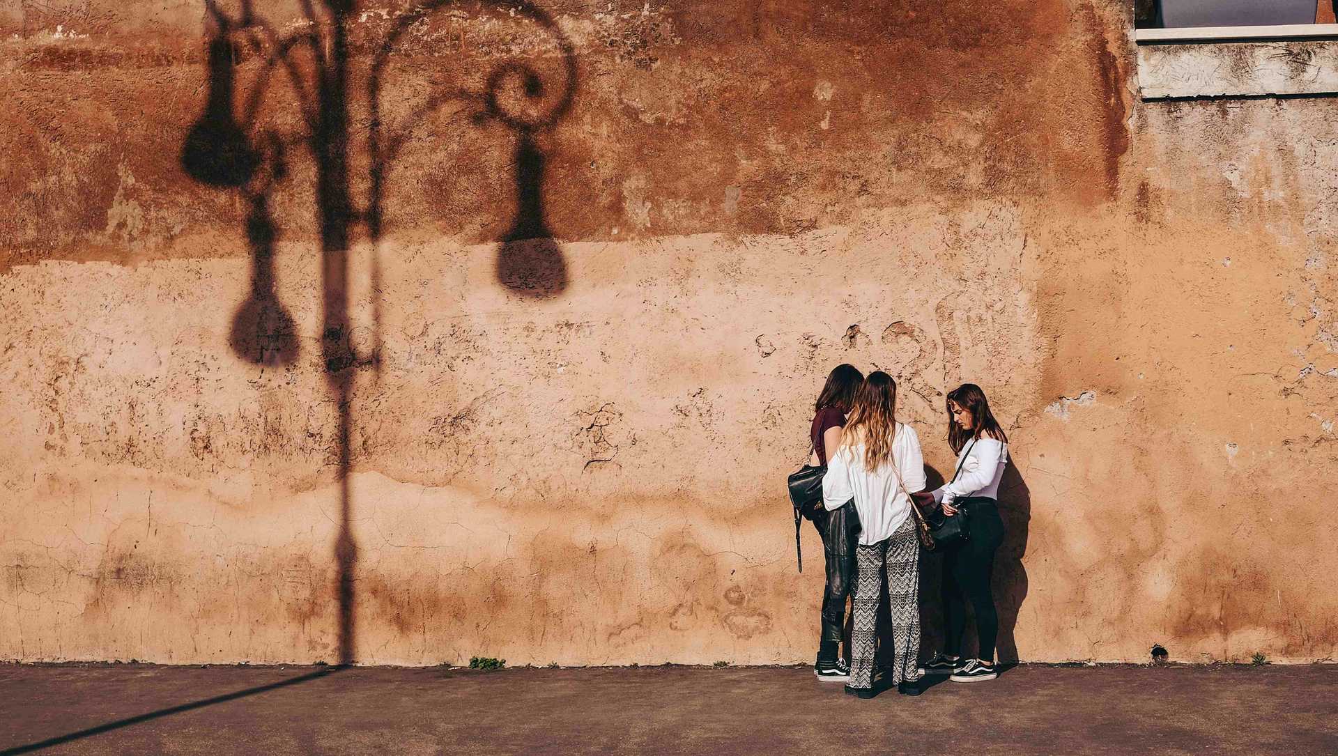 three young women are standing in front of a sunny wall and are talking to each other