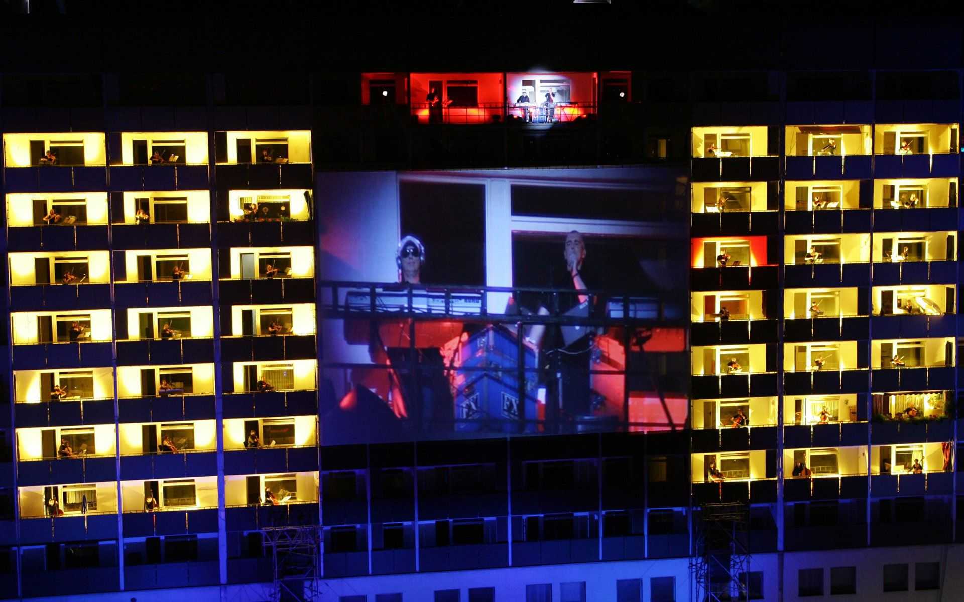 Auf den Balkonen eines Plattenbaus stehen verteilt und hell beleuchtet die Mitglieder der Dresdner Sinfoniker und spielen ihre Instrumente. In der Mitte hängt eine große Leinwand auf der die Übertragung der Sinfonie läuft.
