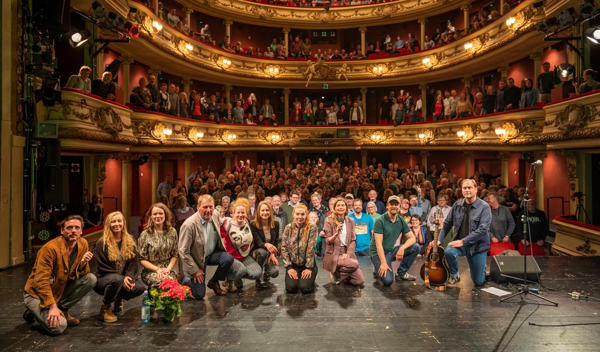 Die Crew der Roadshow  hockt auf der Bühne vor dem Publikum in Oldenburger Staatstheater. Der Saal ist voller Menschen, alle stehen.