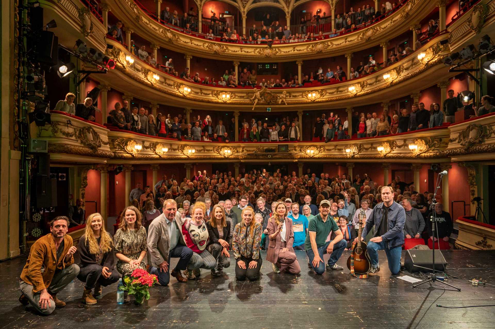 Die Crew der Roadshow  hockt auf der Bühne vor dem Publikum in Oldenburger Staatstheater. Der Saal ist voller Menschen, alle stehen.