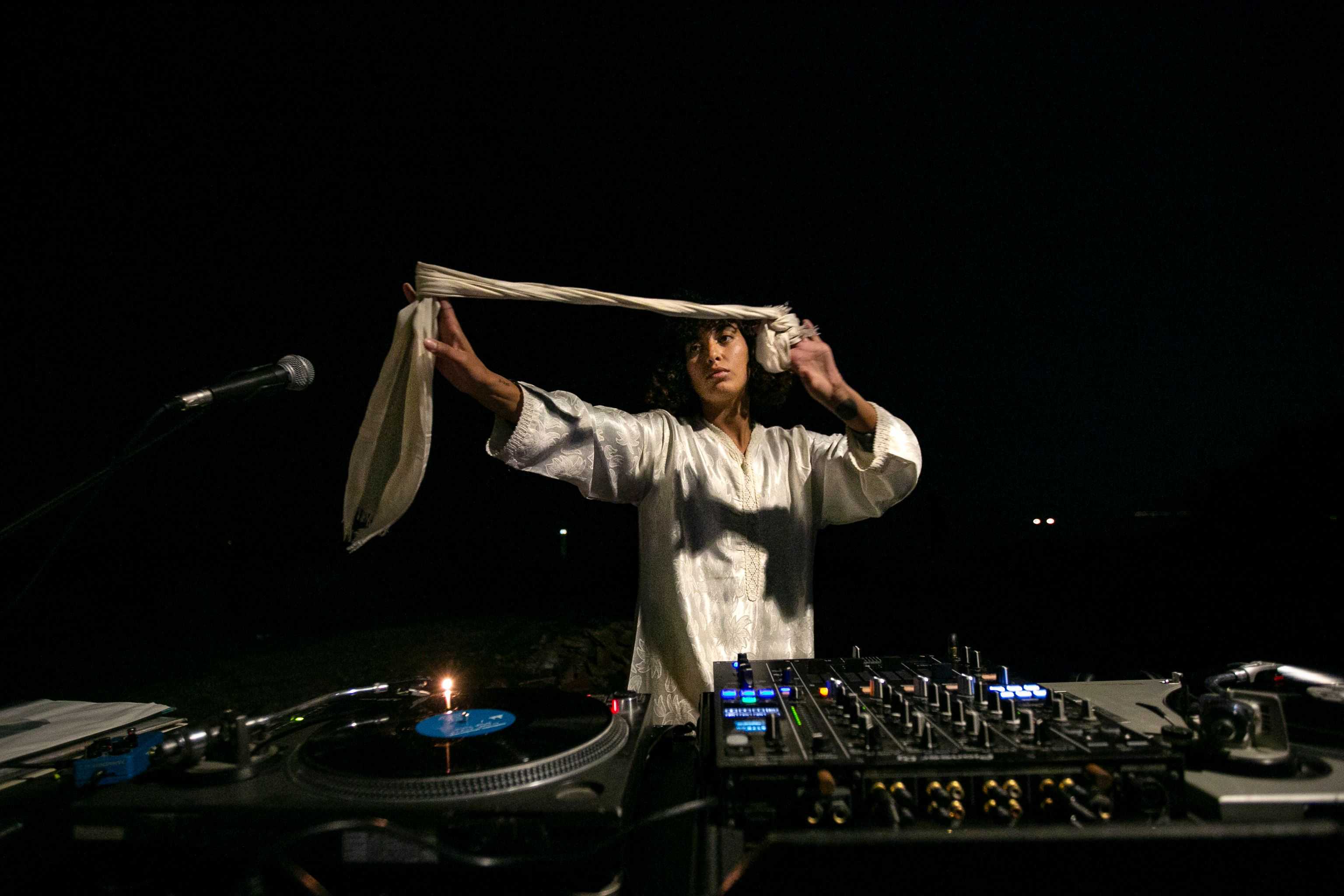 The artist Leila Bencharnia is playing at DJdesks in the nighttime and holds a scarve in her hands