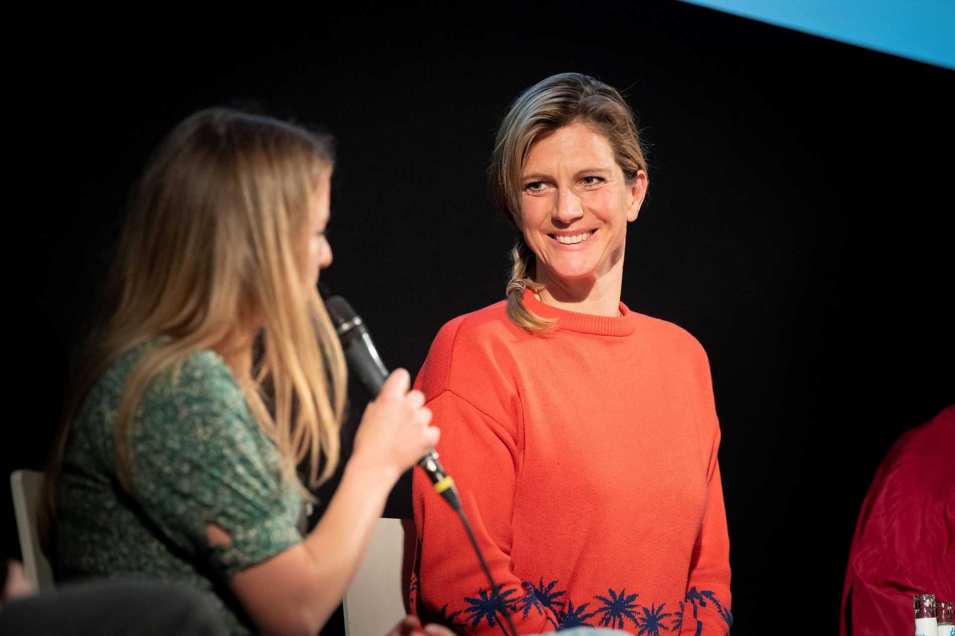 Maja Göpel lächelt eine Person an, die ein Mikrofon in der Hand hält und reinspricht. Sie scheint sich an Maja Göpel zu wenden.