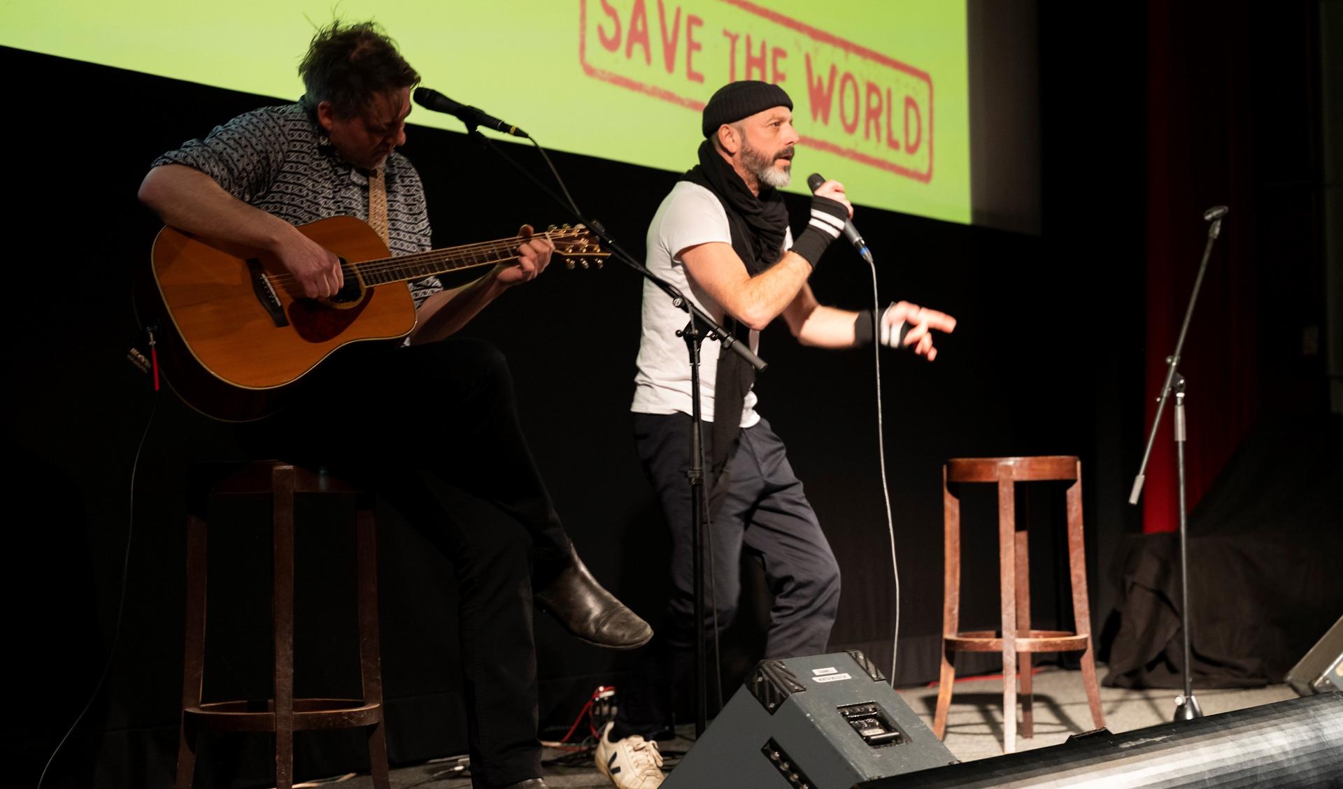 Zwei Musizierende sind auf der Bühne. Die eine Person singt in ein Mikrofon und steht im Bühnenlicht, die andere sitzt auf einem Hocker und spielt eine Gitarre