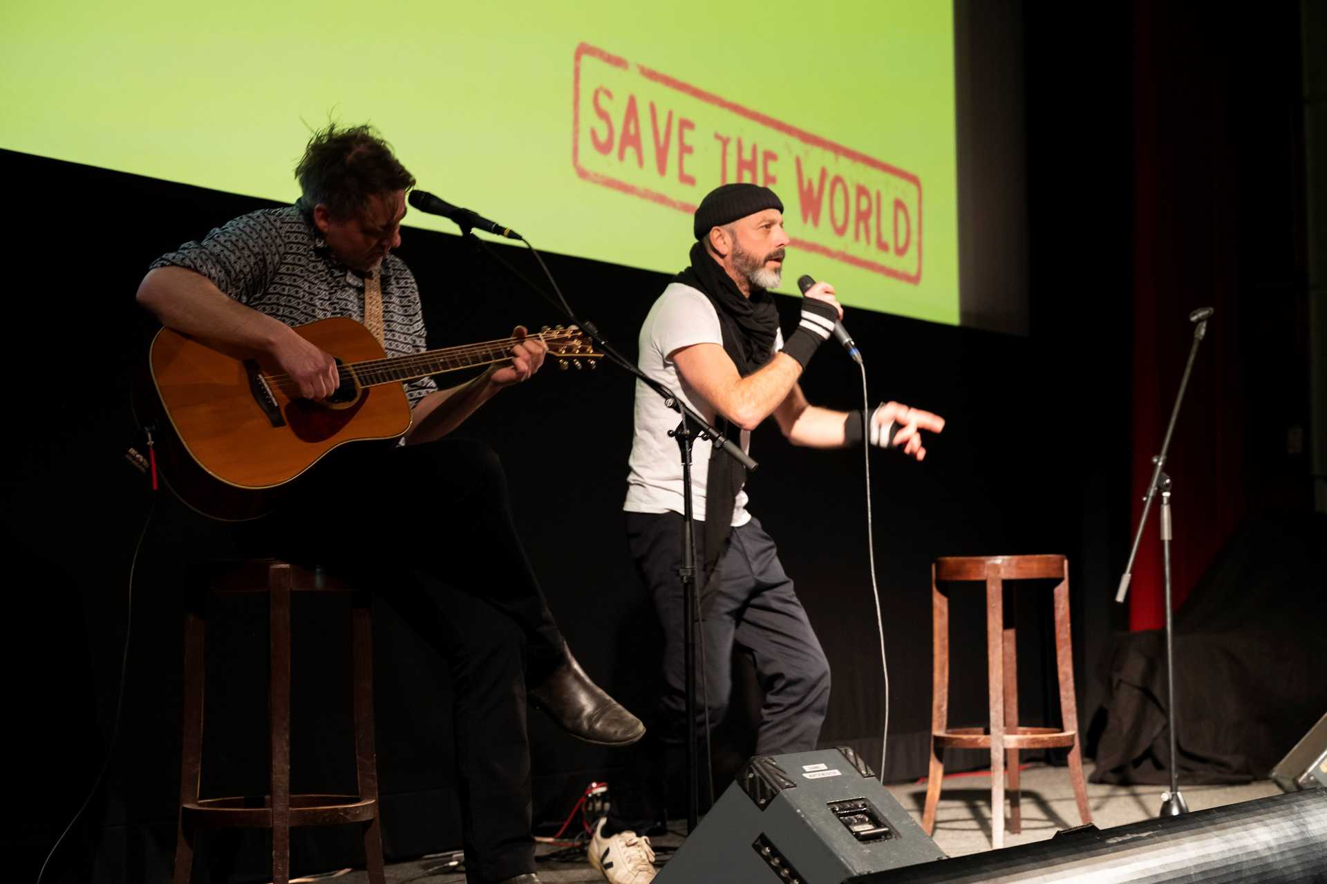 Zwei Musizierende sind auf der Bühne. Die eine Person singt in ein Mikrofon und steht im Bühnenlicht, die andere sitzt auf einem Hocker und spielt eine Gitarre