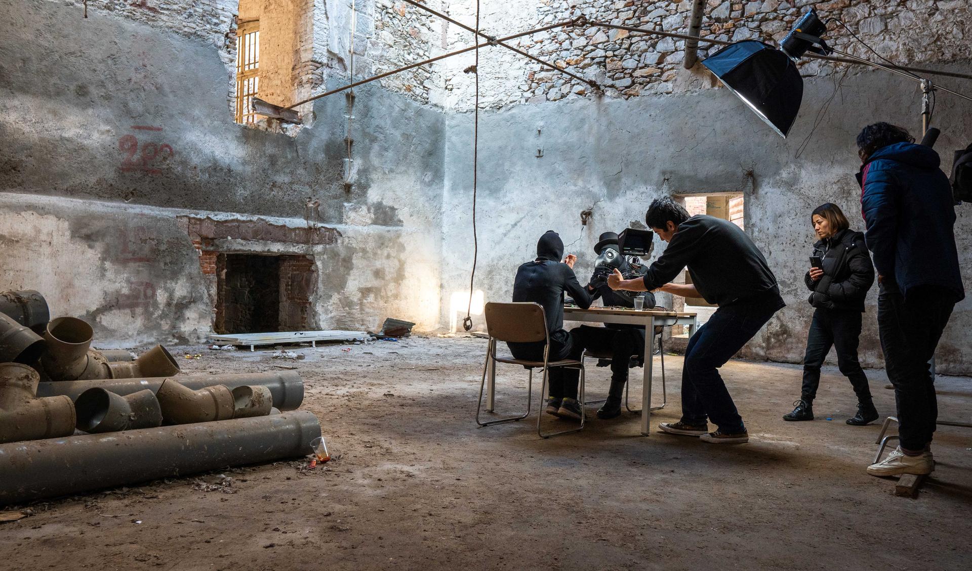 Five persons in a room are making a movie. Two are sitting at a table in a shabby and cold room and the others are filming them.