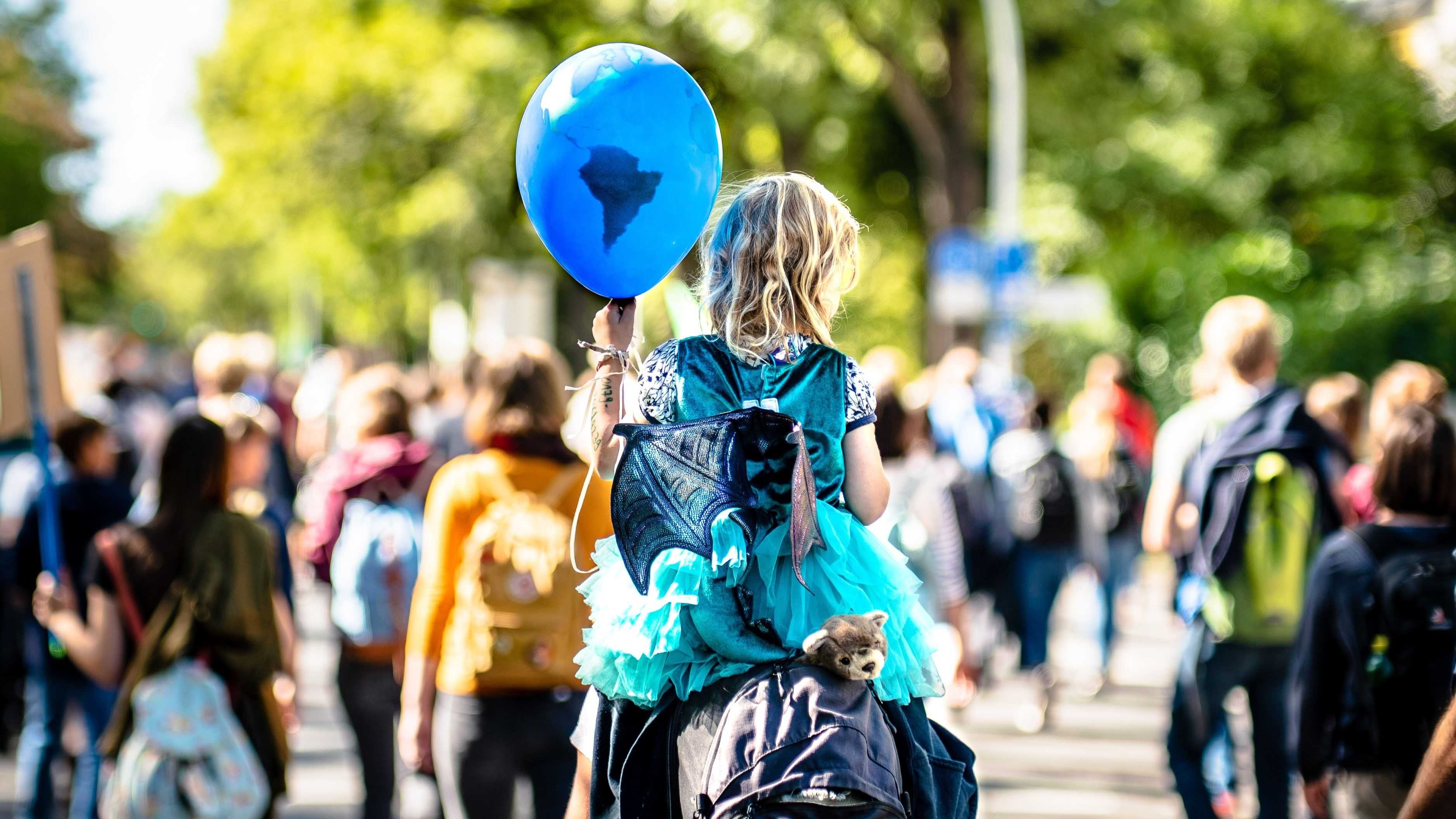 A demonstration - a child is sitting on the shoulders of a grown-uo person. It holds a ballon in its hands that looks like the earth but in blue.