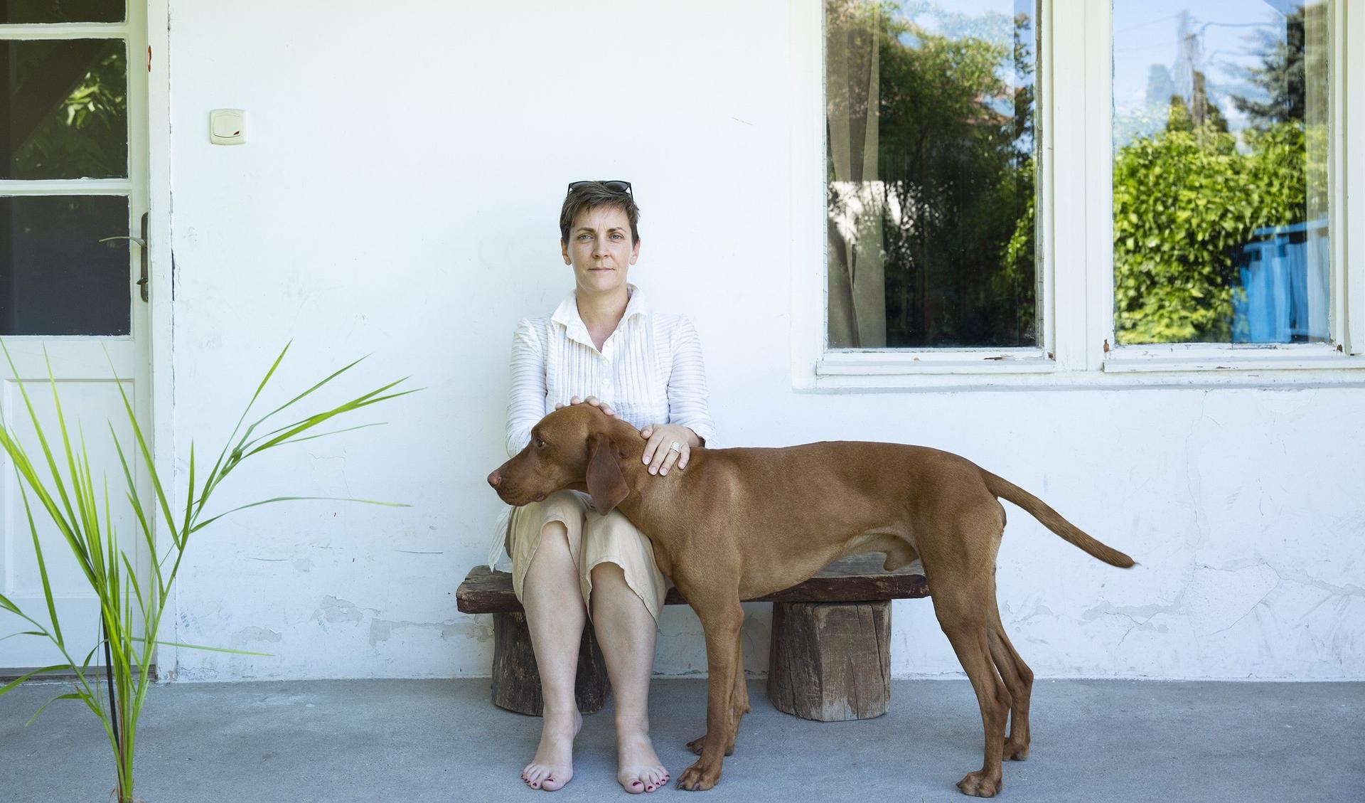 Kata Csato is sitting in fron of a white house on a bench and is looking into the camera. She is petting a big brown dog.