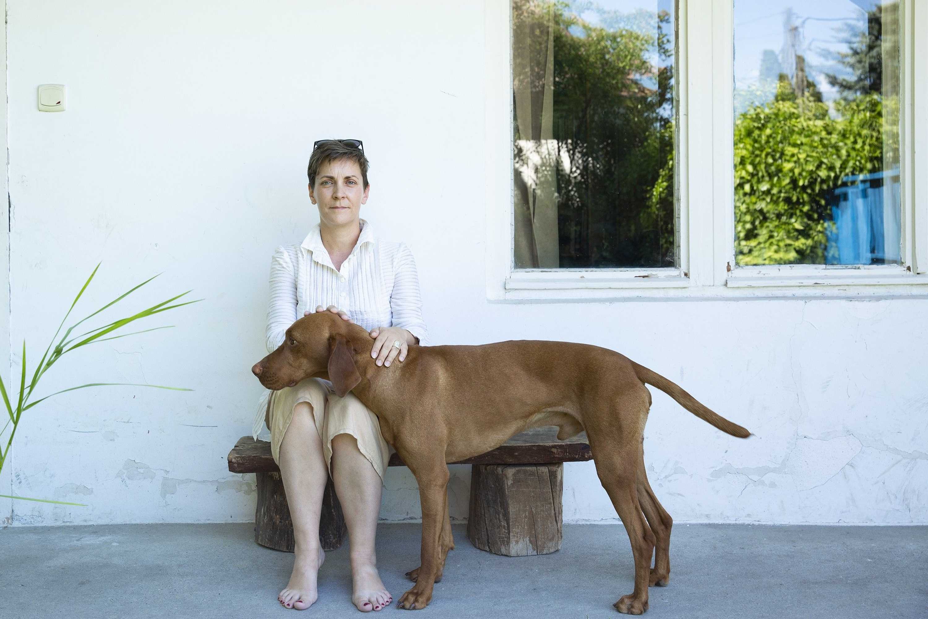 Kata Csato is sitting in fron of a white house on a bench and is looking into the camera. She is petting a big brown dog.