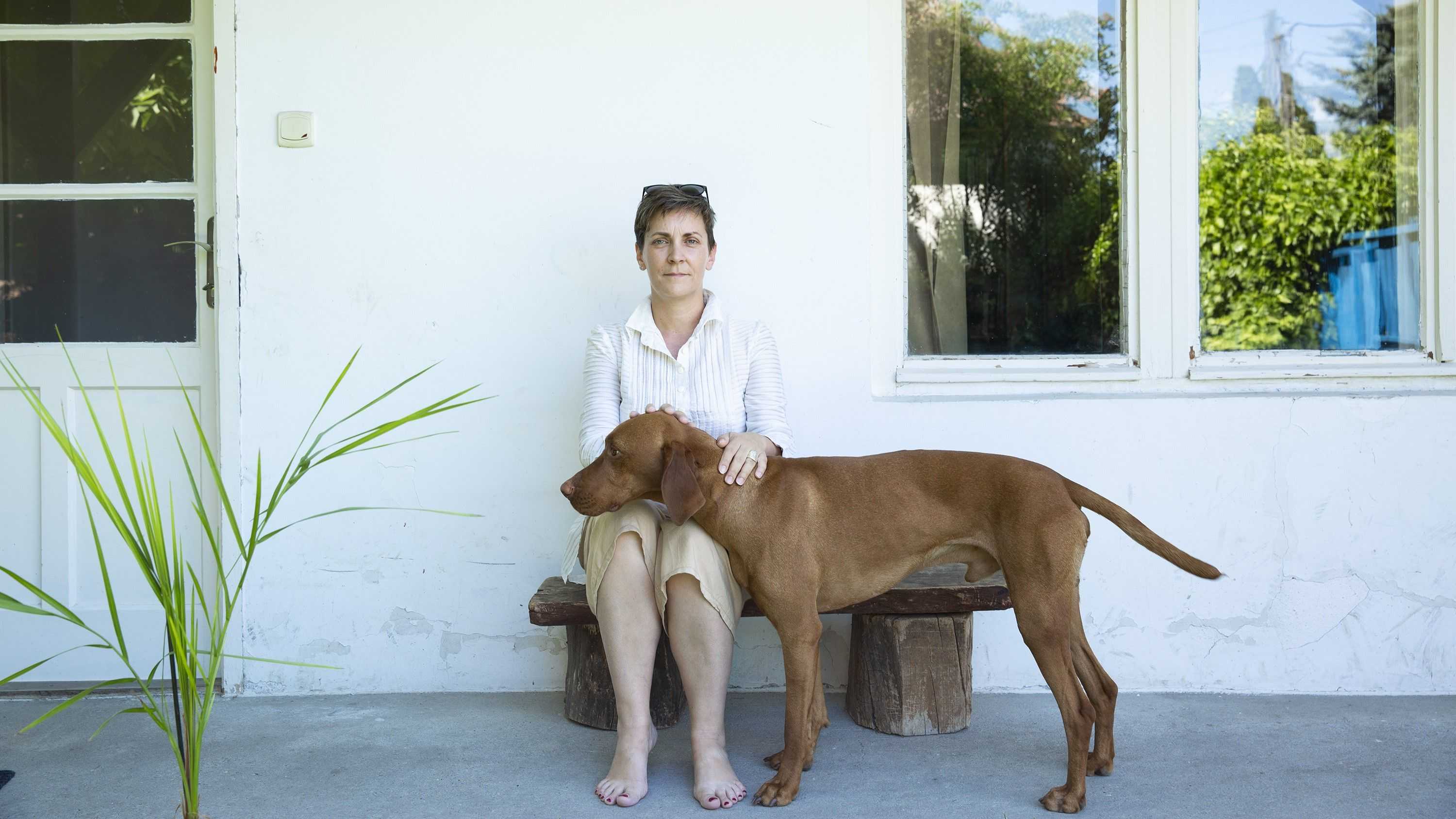 Kata Csato is sitting in fron of a white house on a bench and is looking into the camera. She is petting a big brown dog.