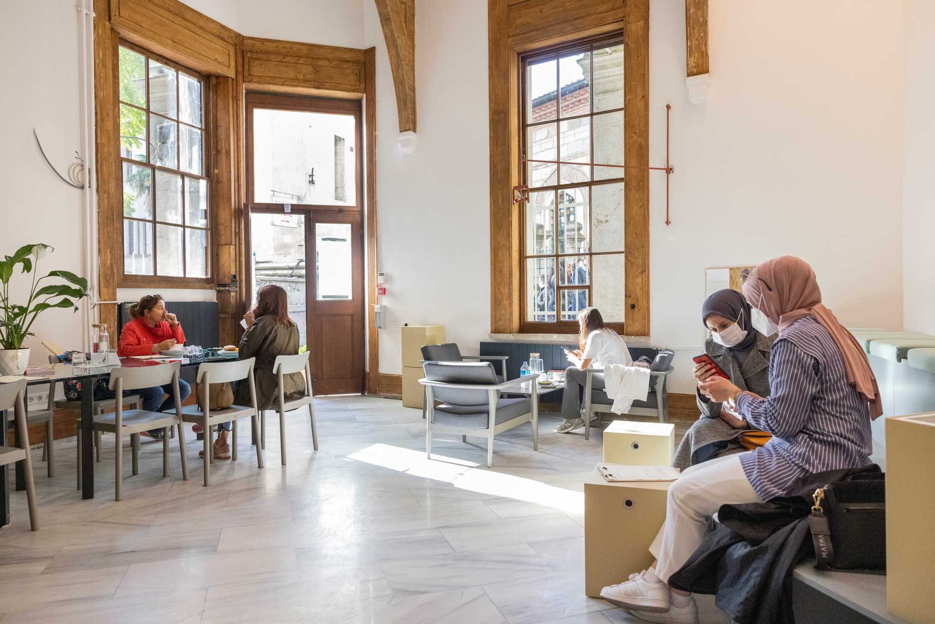 People are sitting in a sunny room and are chatting and working on their laptops.