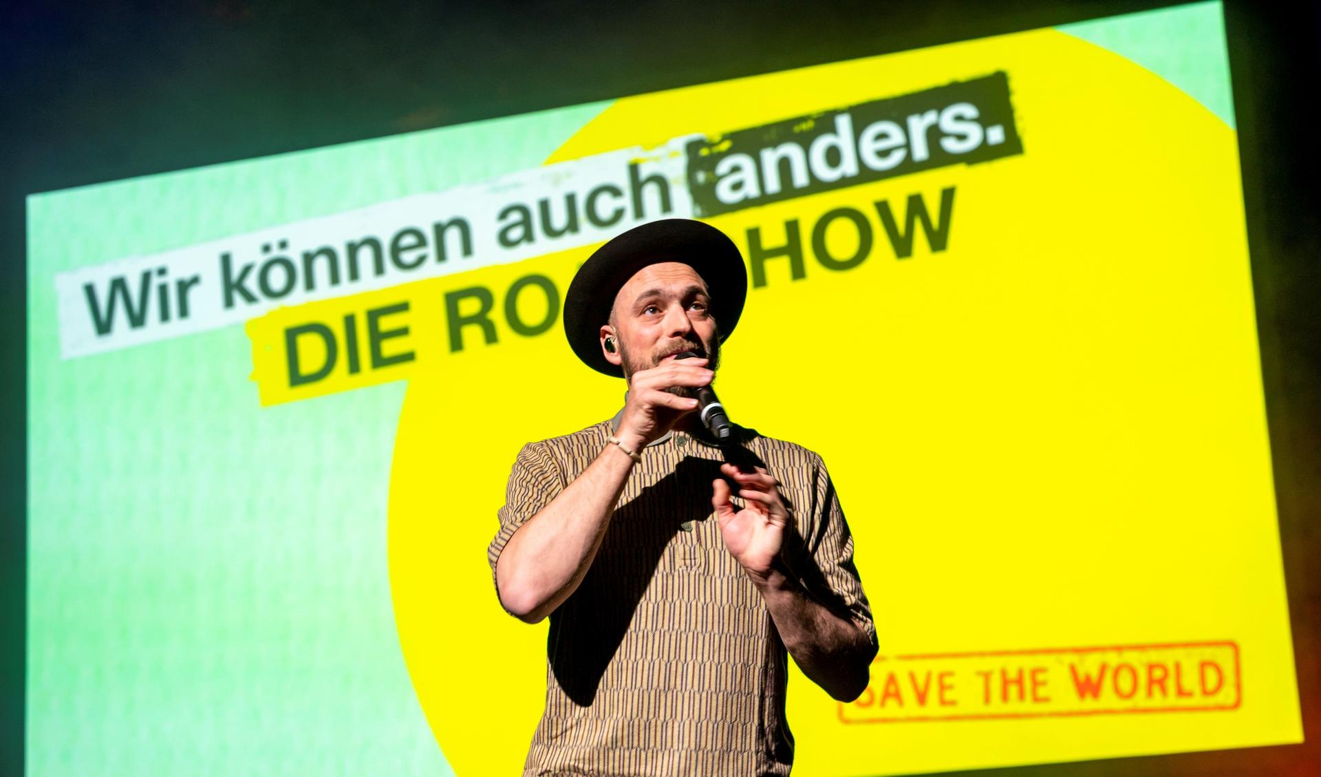 Eine Person mit Hut und einem Mikrofon in der Hand singt auf einer Bühhne. Sie steht vor einer Leinwand mit dem ogo der Roadshow.