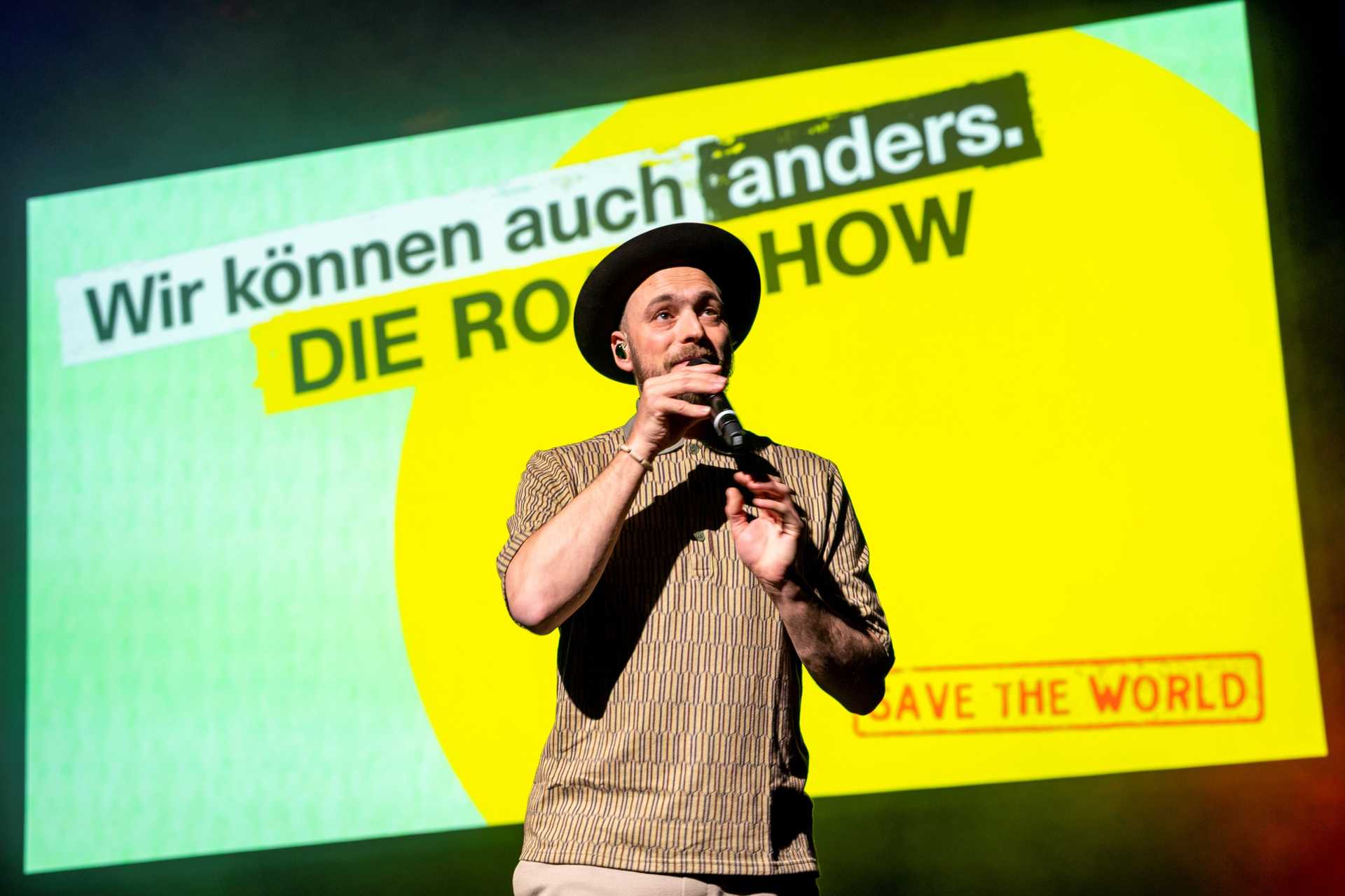 Eine Person mit Hut und einem Mikrofon in der Hand singt auf einer Bühhne. Sie steht vor einer Leinwand mit dem ogo der Roadshow.