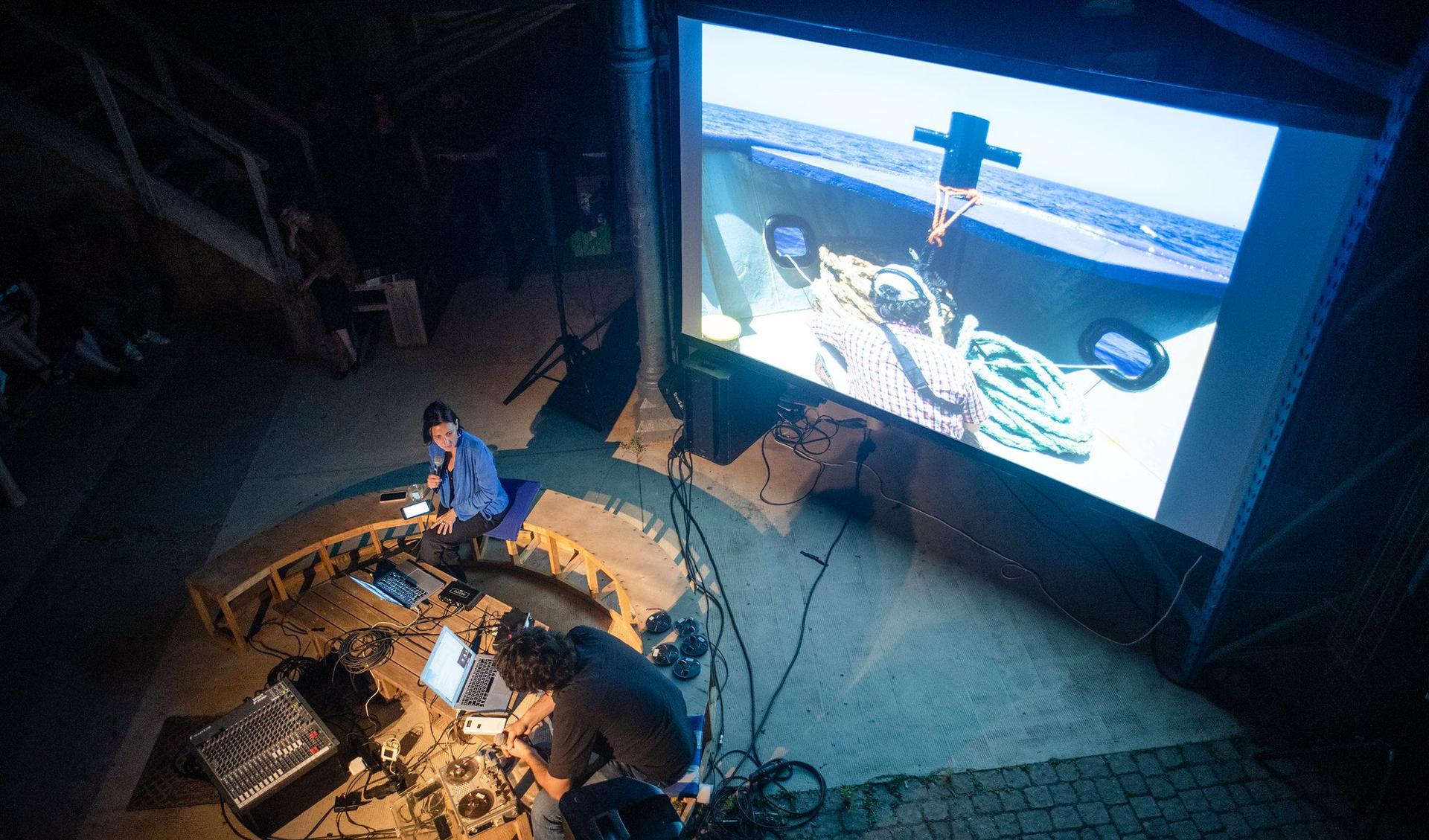 The photo looks from the top on two people sitting on a bench in front of their laptops and some equipment- In the background a movie is playing.