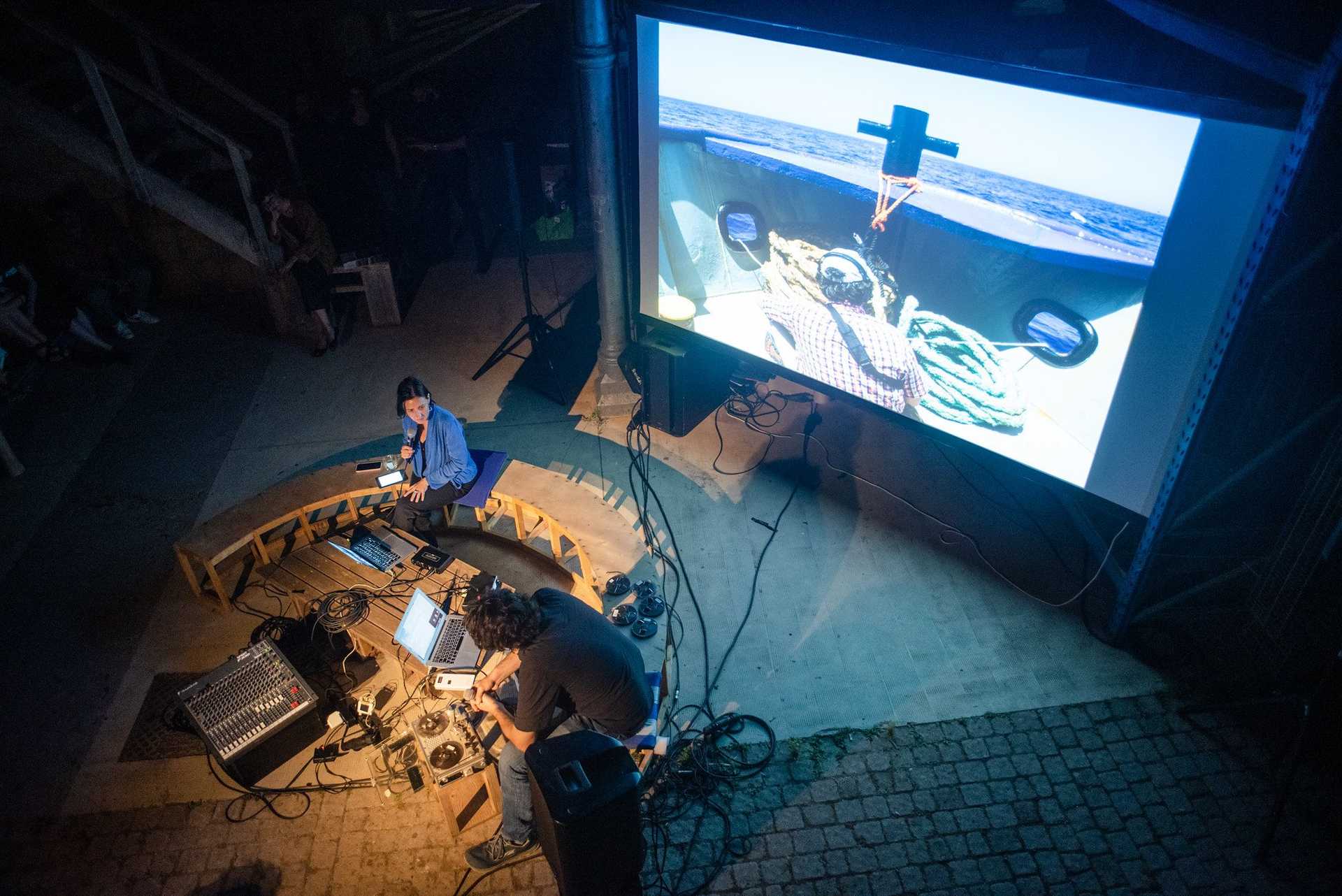 The photo looks from the top on two people sitting on a bench in front of their laptops and some equipment- In the background a movie is playing.