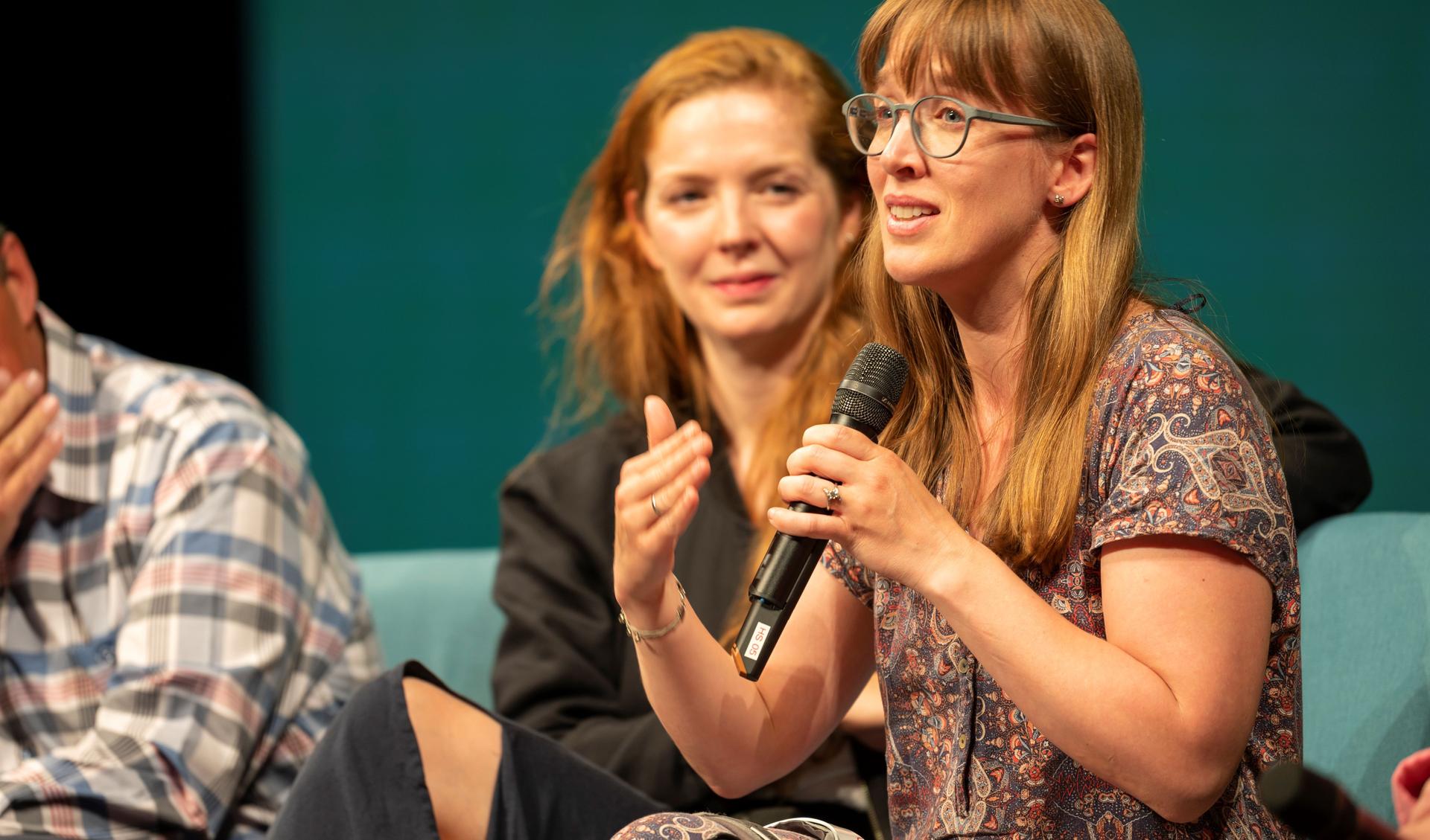 A person is speaking to the audience with a microphone sitting on a chair. Another person sits next to her and is smiling at her and listening