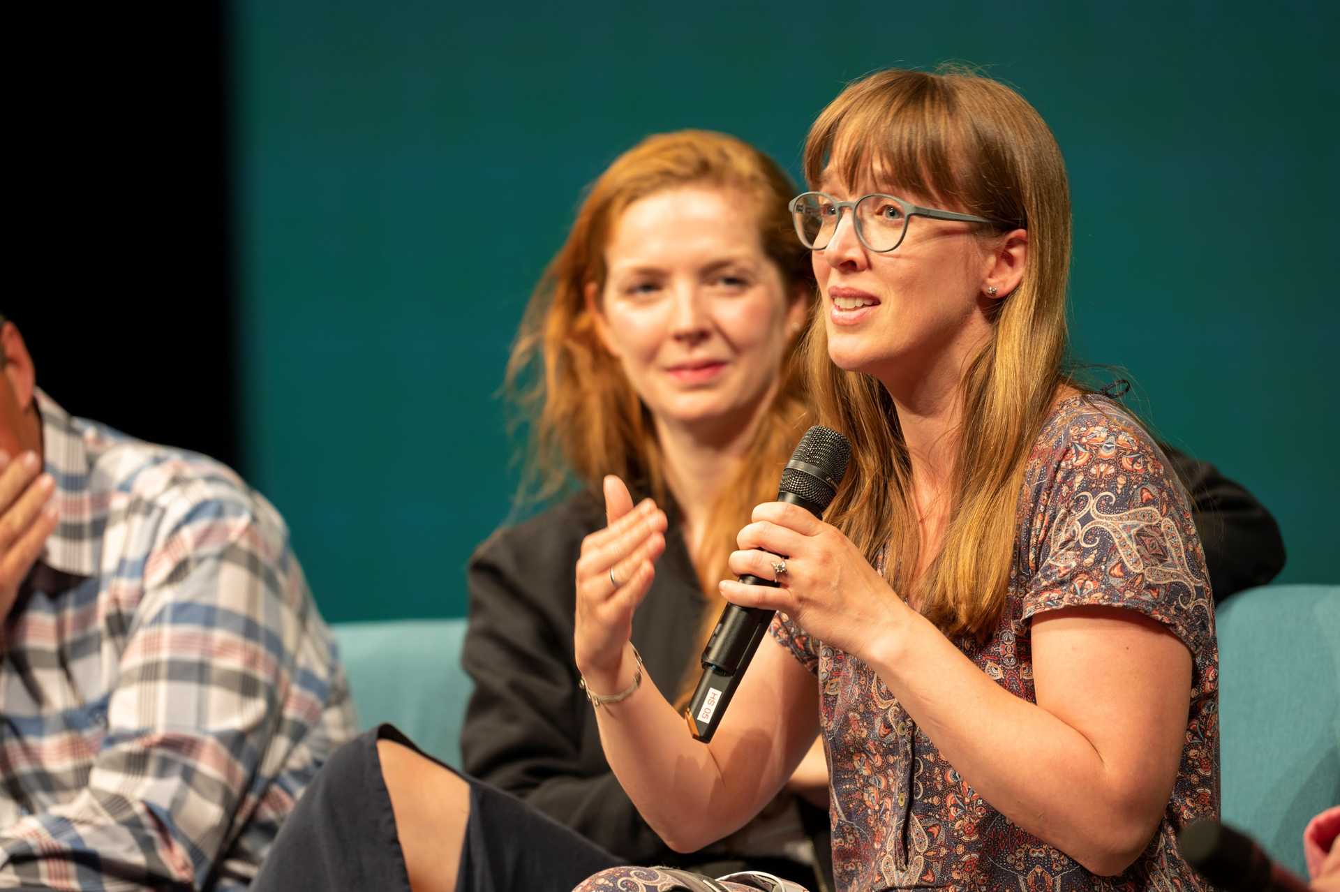 A person is speaking to the audience with a microphone sitting on a chair. Another person sits next to her and is smiling at her and listening