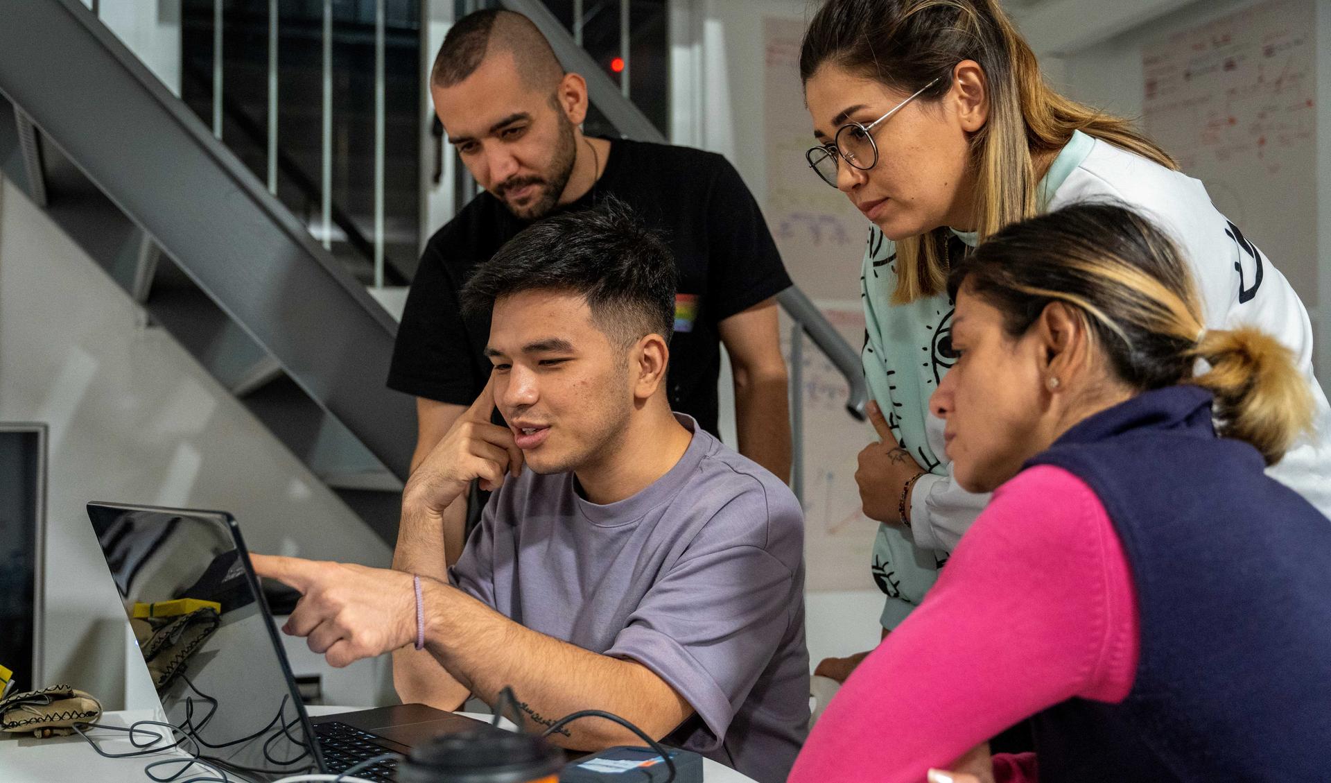 A person is showing three others something on a laptop. The other three are looking very focussed.