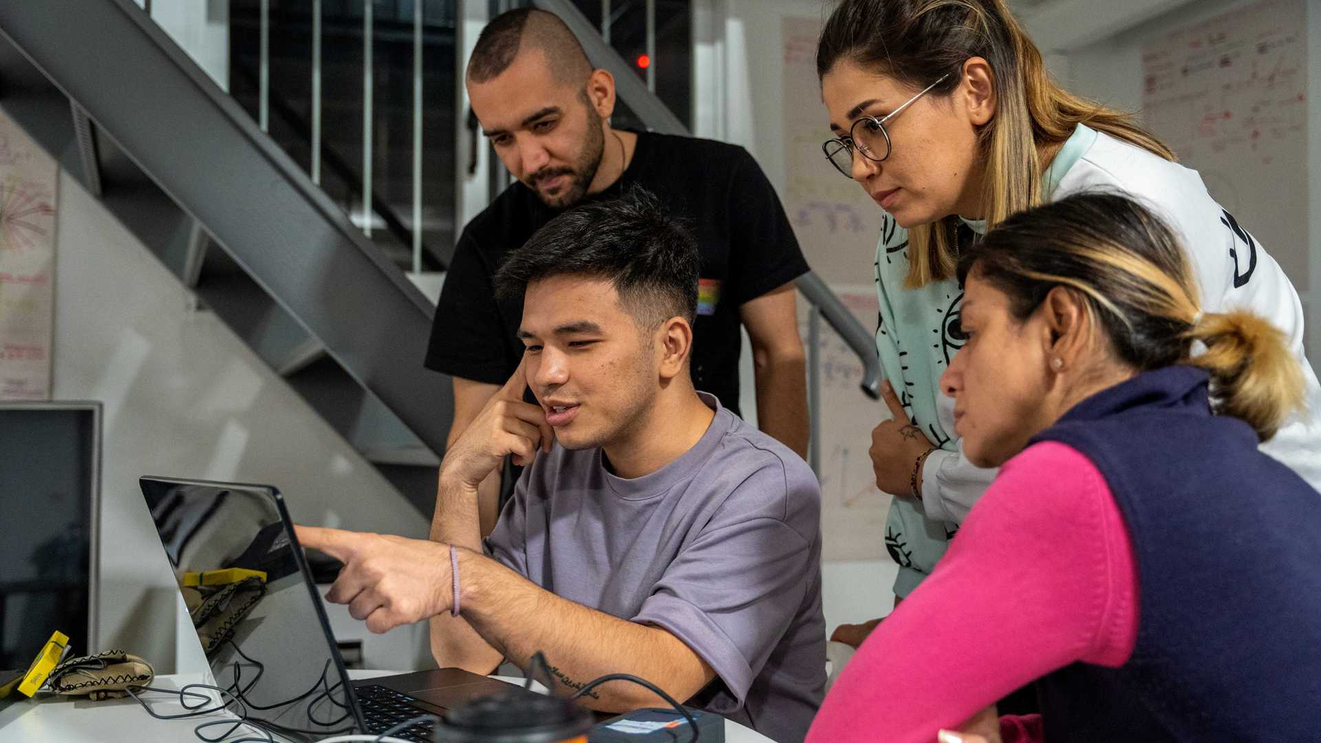 A person is showing three others something on a laptop. The other three are looking very focussed.
