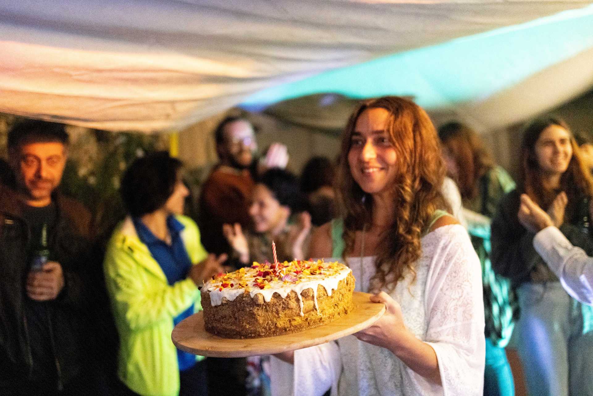 A person is carrying a birthday cake in a room that is full with people.