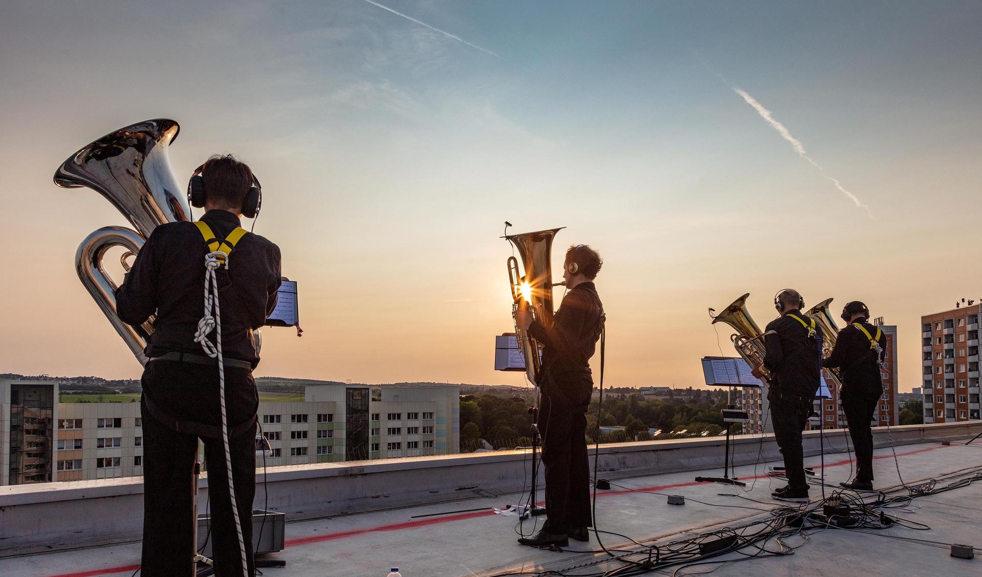 Four blowers, members of the Dresdner Sinfoniker are standing on the roof o a tower-builidng and are playing their instruments. They are secures with climbing gear and the sun goes down in the background