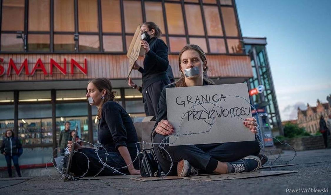 Aleksandra Łoboda is sitting on a street, her mouth taped with gaffa and around her and two colleagues there is barbed wire. She is holding a sign that is saying: The border of Decency