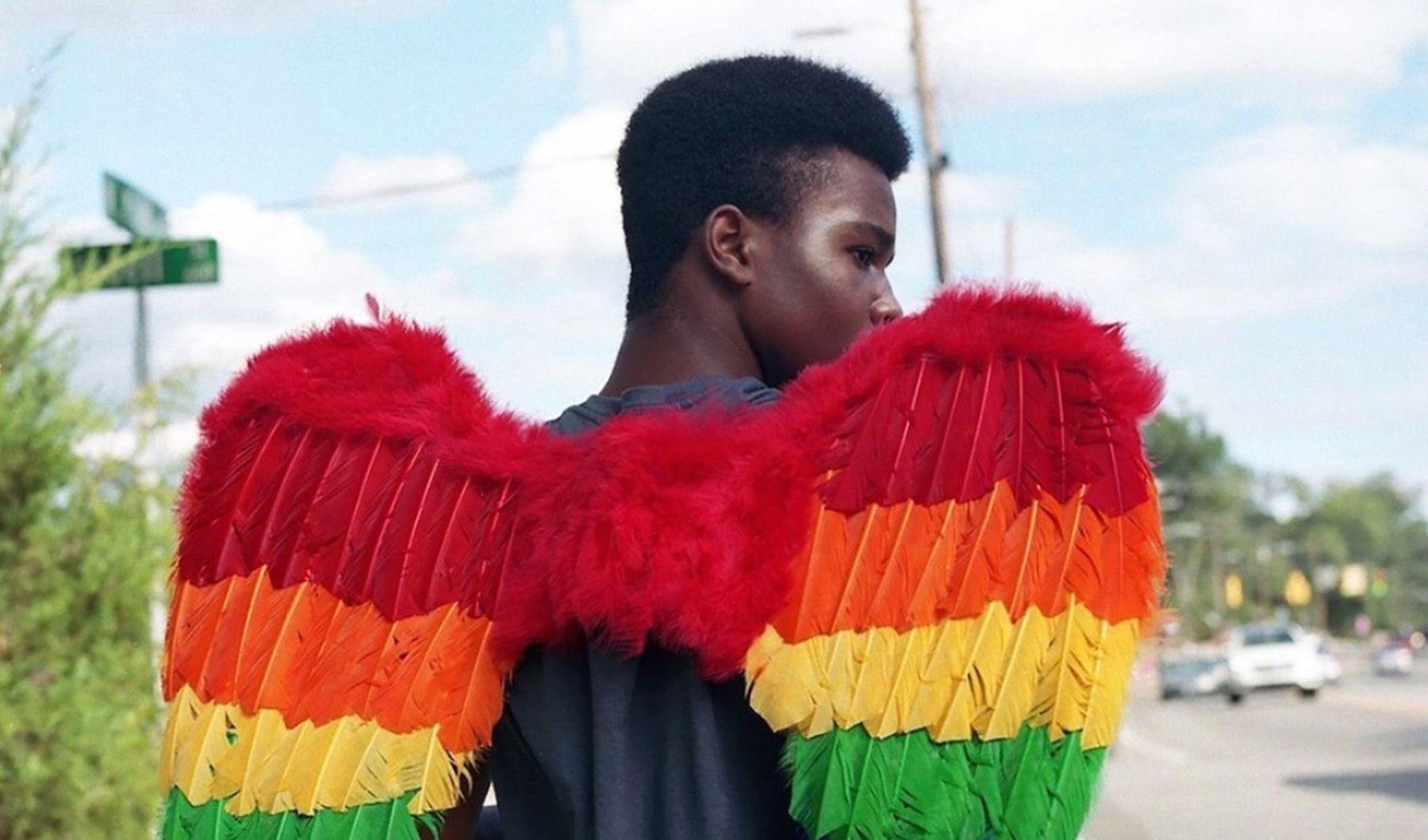 Eine Person mit Engelsflügeln in Regenbogenfarben geht eine Straße entlang. Die Person ist von hinten zu sehen und dreht ihren Kopf leicht zur Seite.