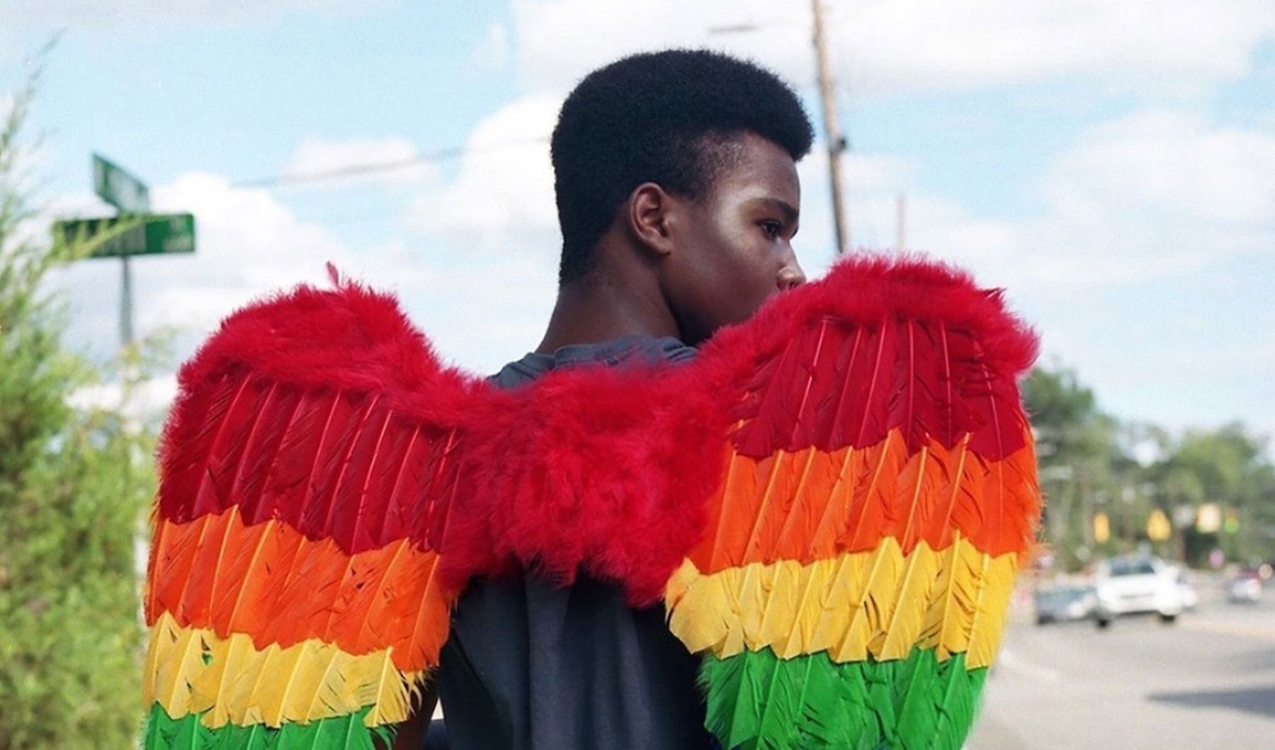 Eine Person mit Engelsflügeln in Regenbogenfarben geht eine Straße entlang. Die Person ist von hinten zu sehen und dreht ihren Kopf leicht zur Seite.