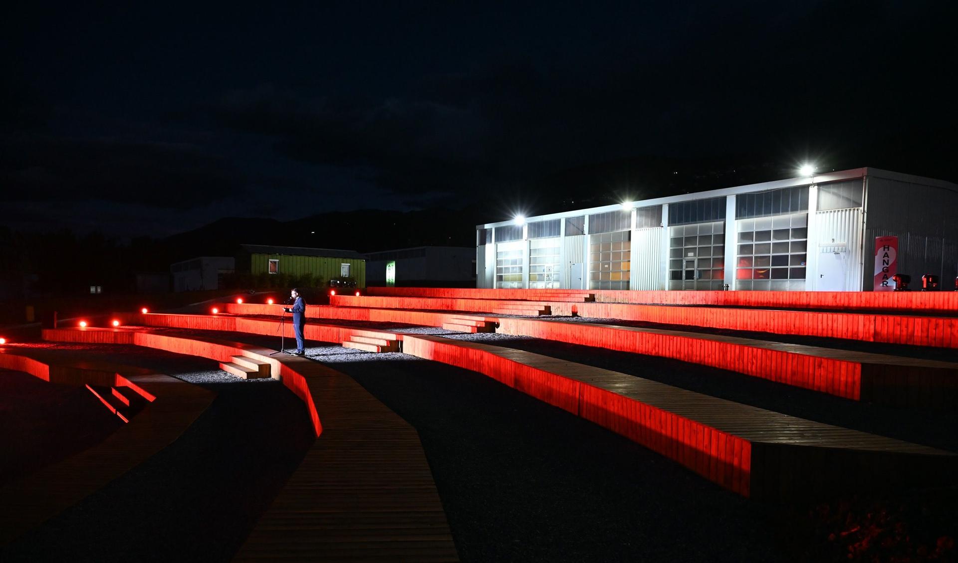 A person is standing, very well dressed in the front of a hangar. Its night time and the person is holding a speech.