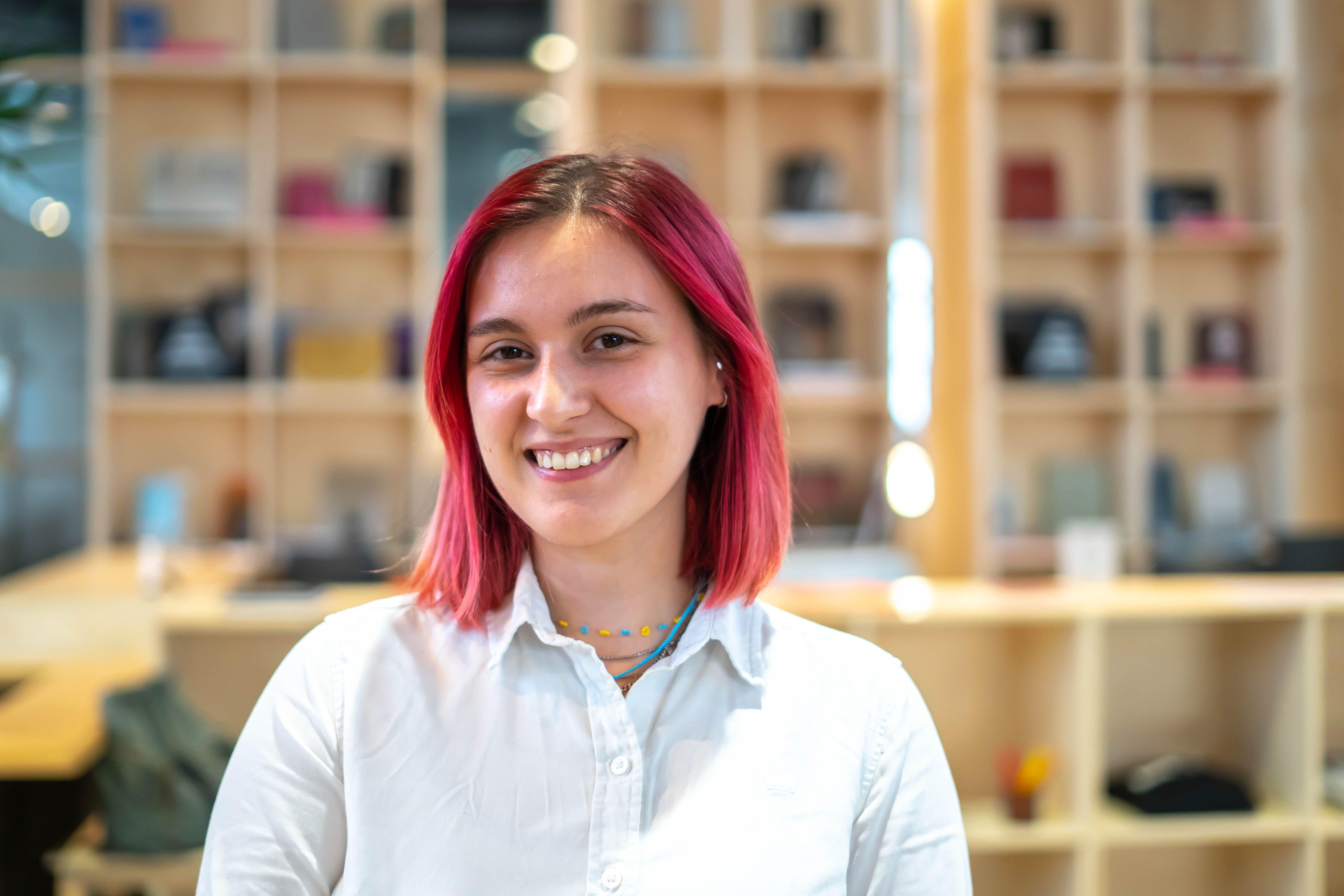 A person with red hair smiles into the camera. The person stands in the middle of the Autostrada Hangar in fron of a shelve with different items in it.